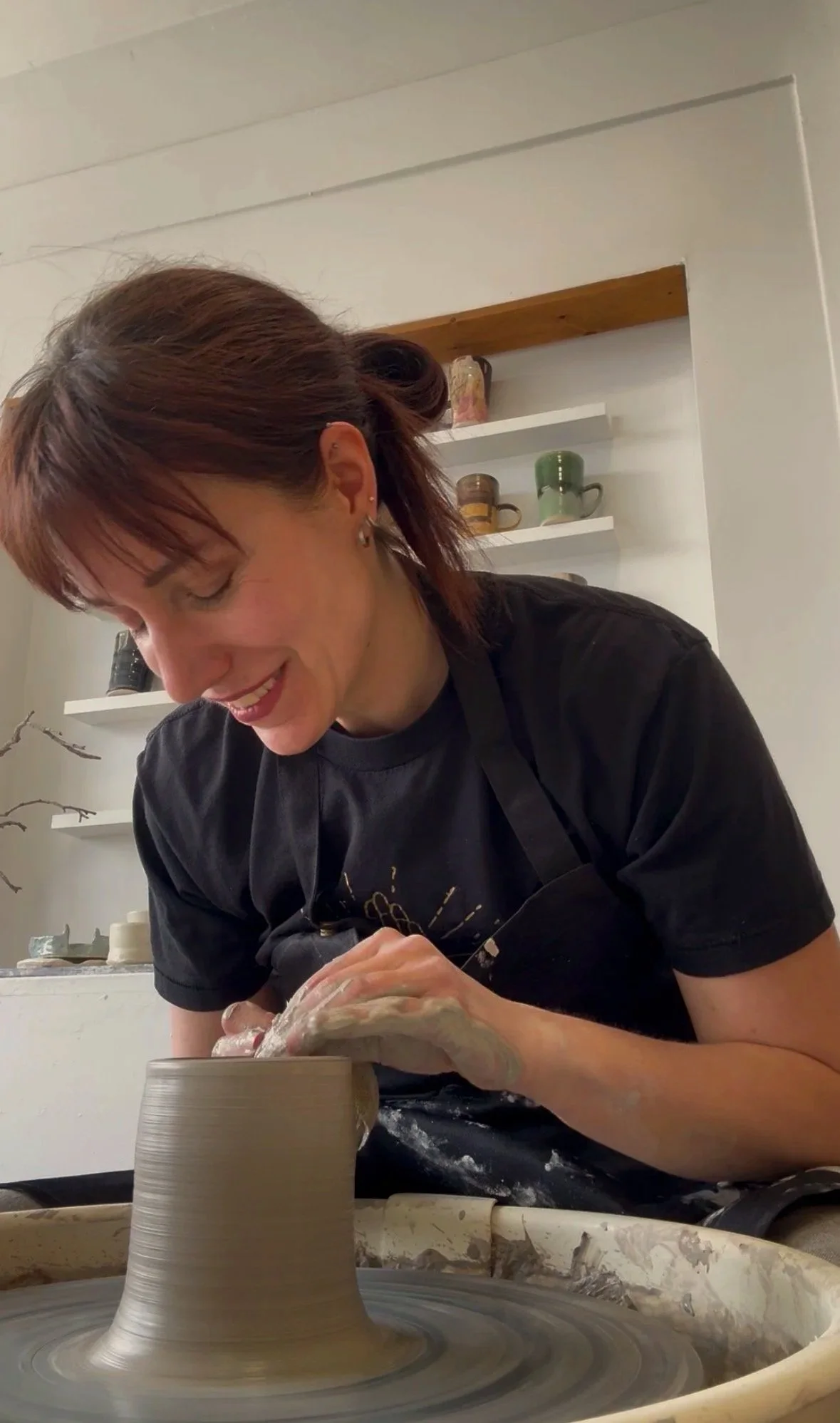 A woman with red hair, wearing a black shirt, is smiling while shaping a ceramic piece on a pottery wheel in a studio.