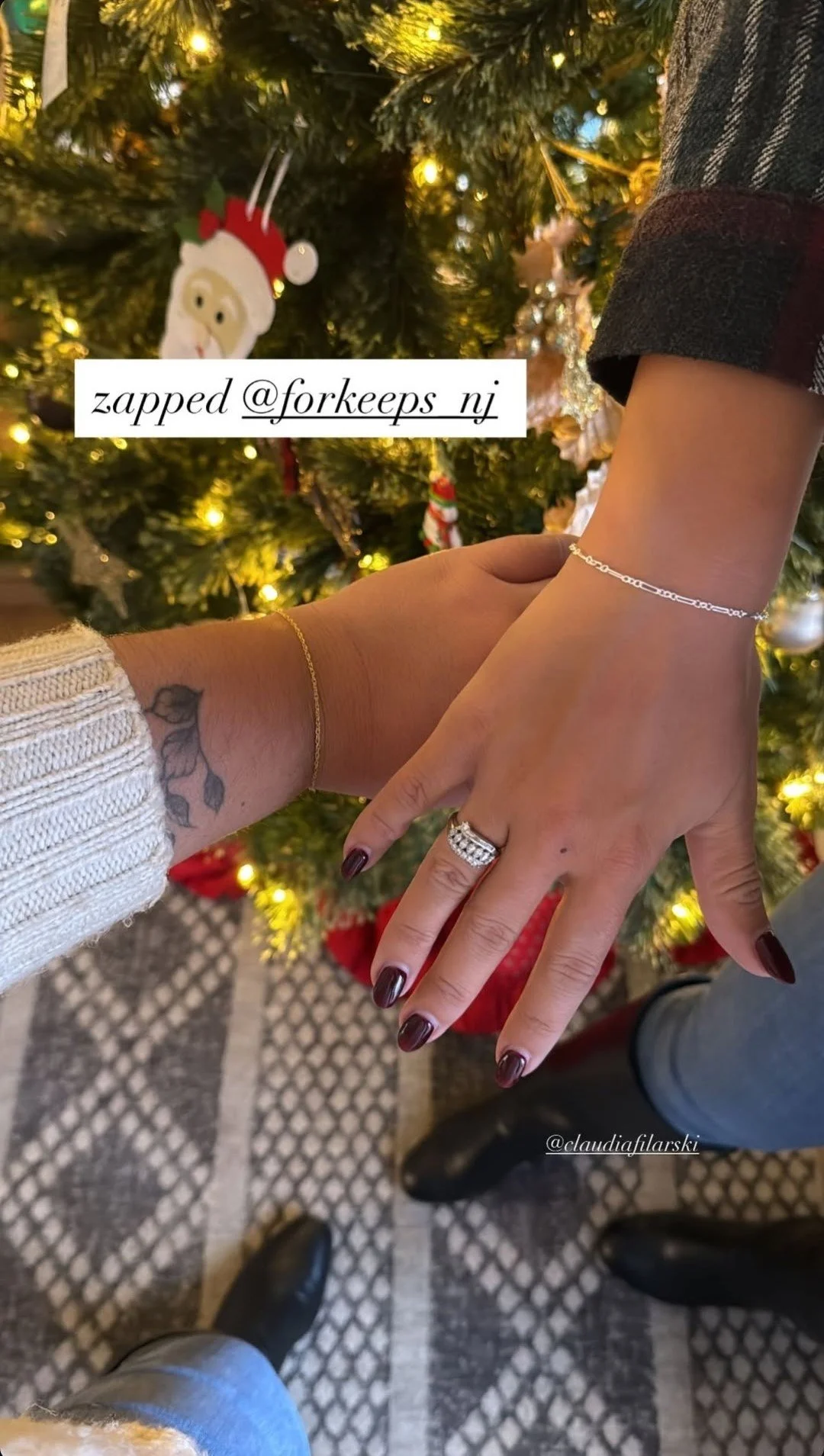 Close-up of two hands showing wedding rings and jewelry with a decorated Christmas tree in the background.
