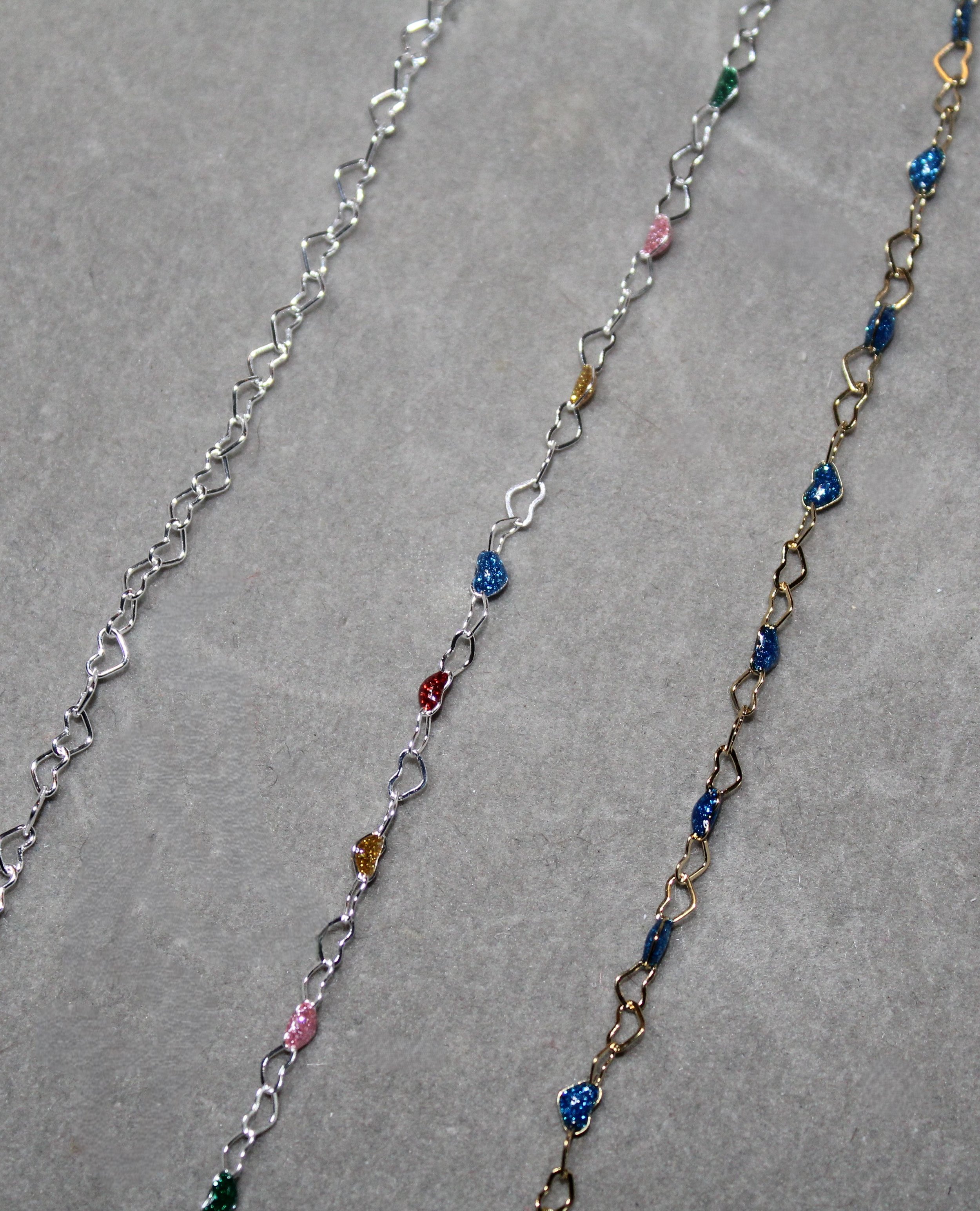 Three necklaces with heart-shaped links and small, sparkling colored beads, displayed on a gray surface.