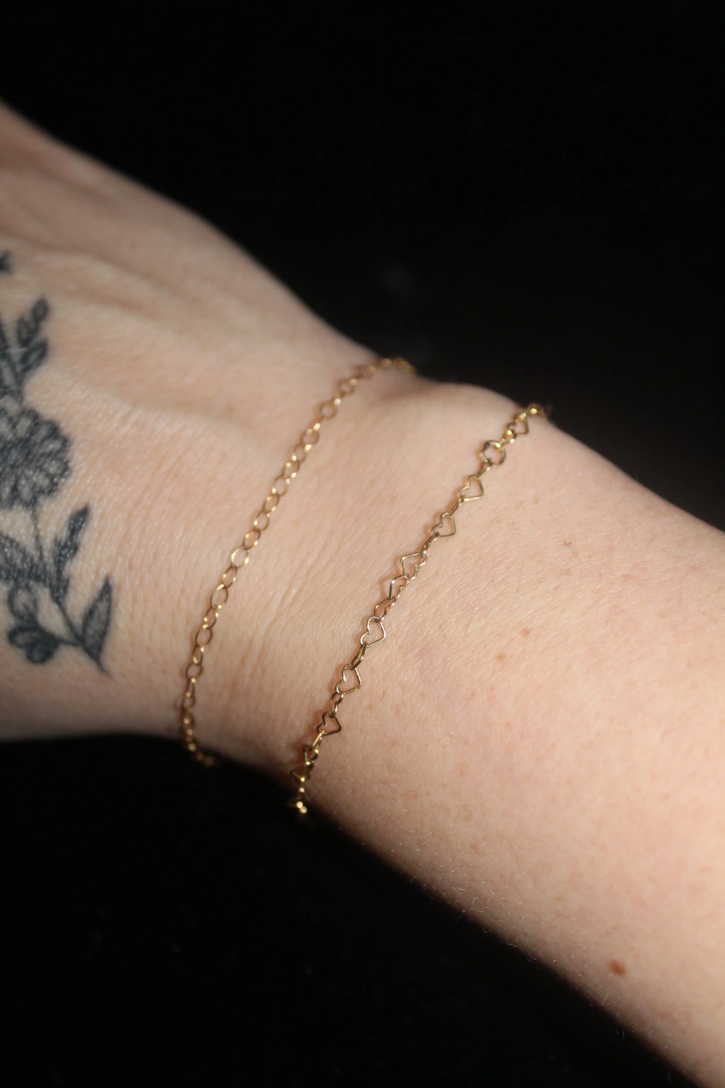 Two delicate gold chain bracelets, one with small heart-shaped links, adorn a wrist with a tattoo of flowers and leaves. The skin is light, and the background is solid black.