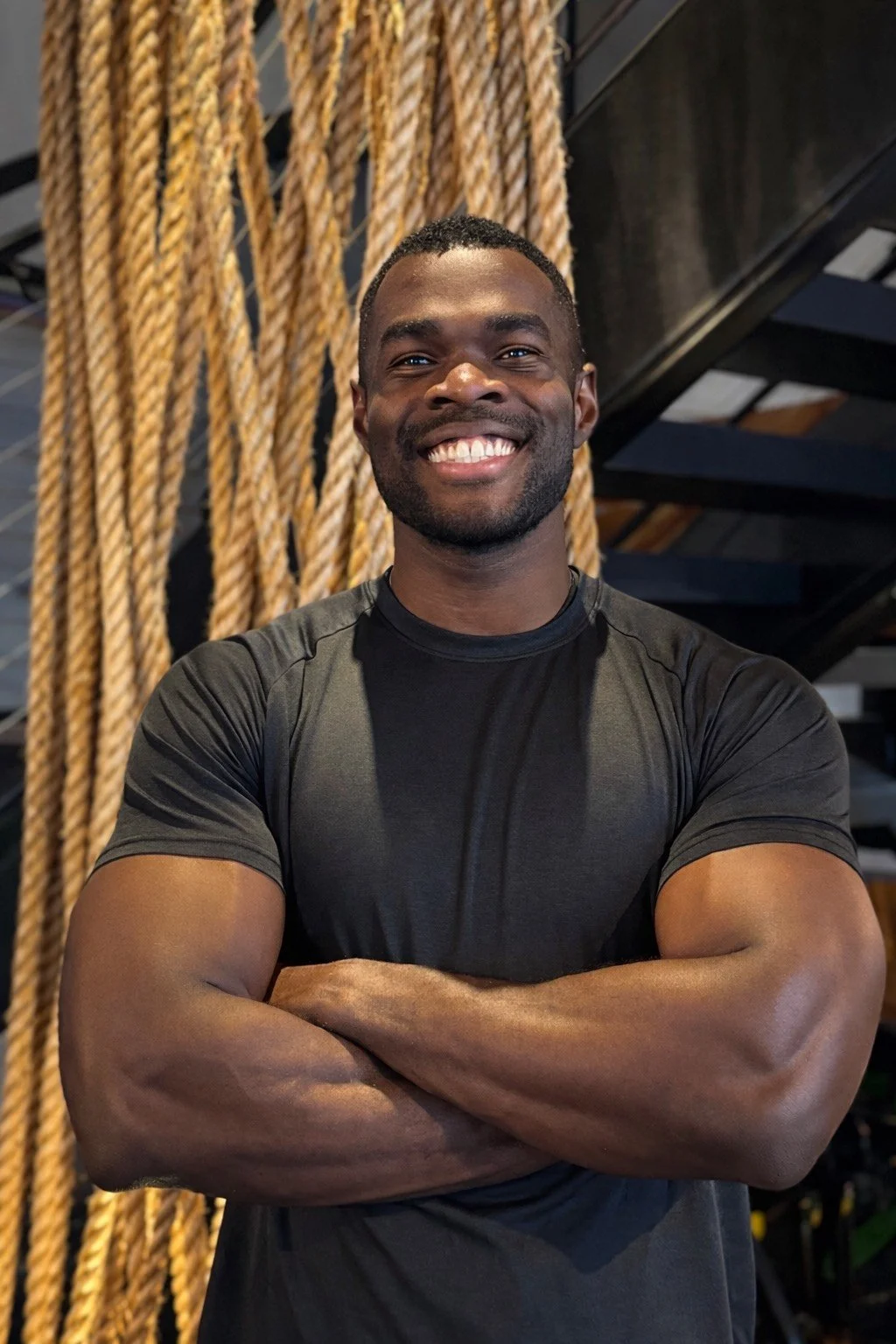 A smiling man with crossed arms wearing a black athletic shirt in a gym with ropes hanging in the background.