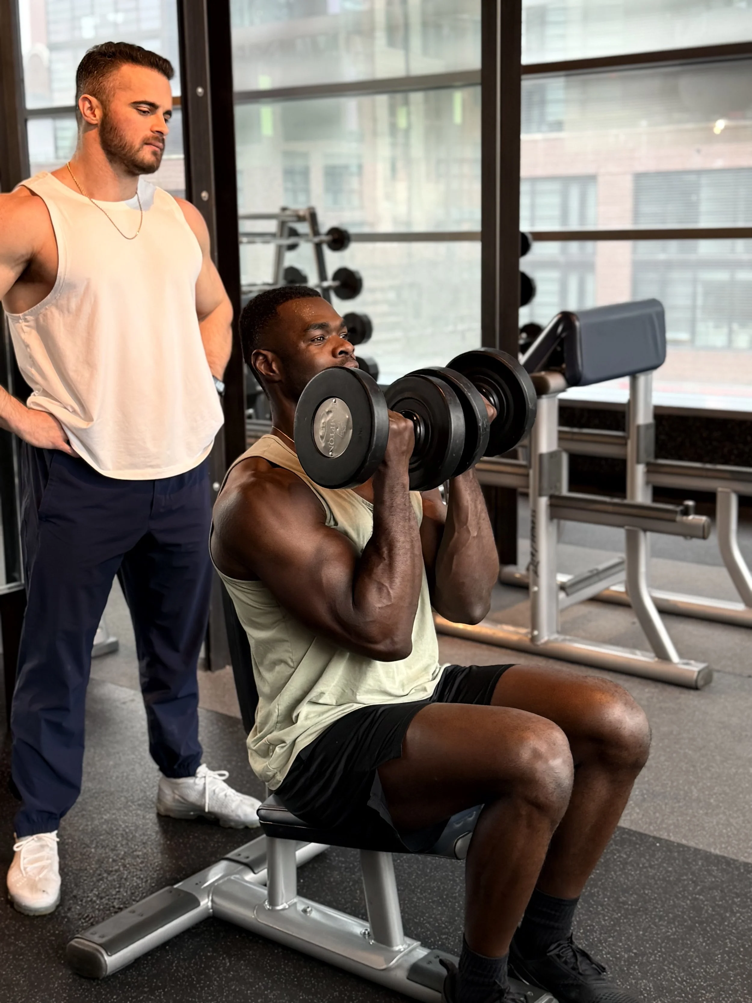 A man sitting on a gym bench performing a seated dumbbell shoulder press, with another man standing behind him observing, in a fitness center with large windows and exercise equipment.