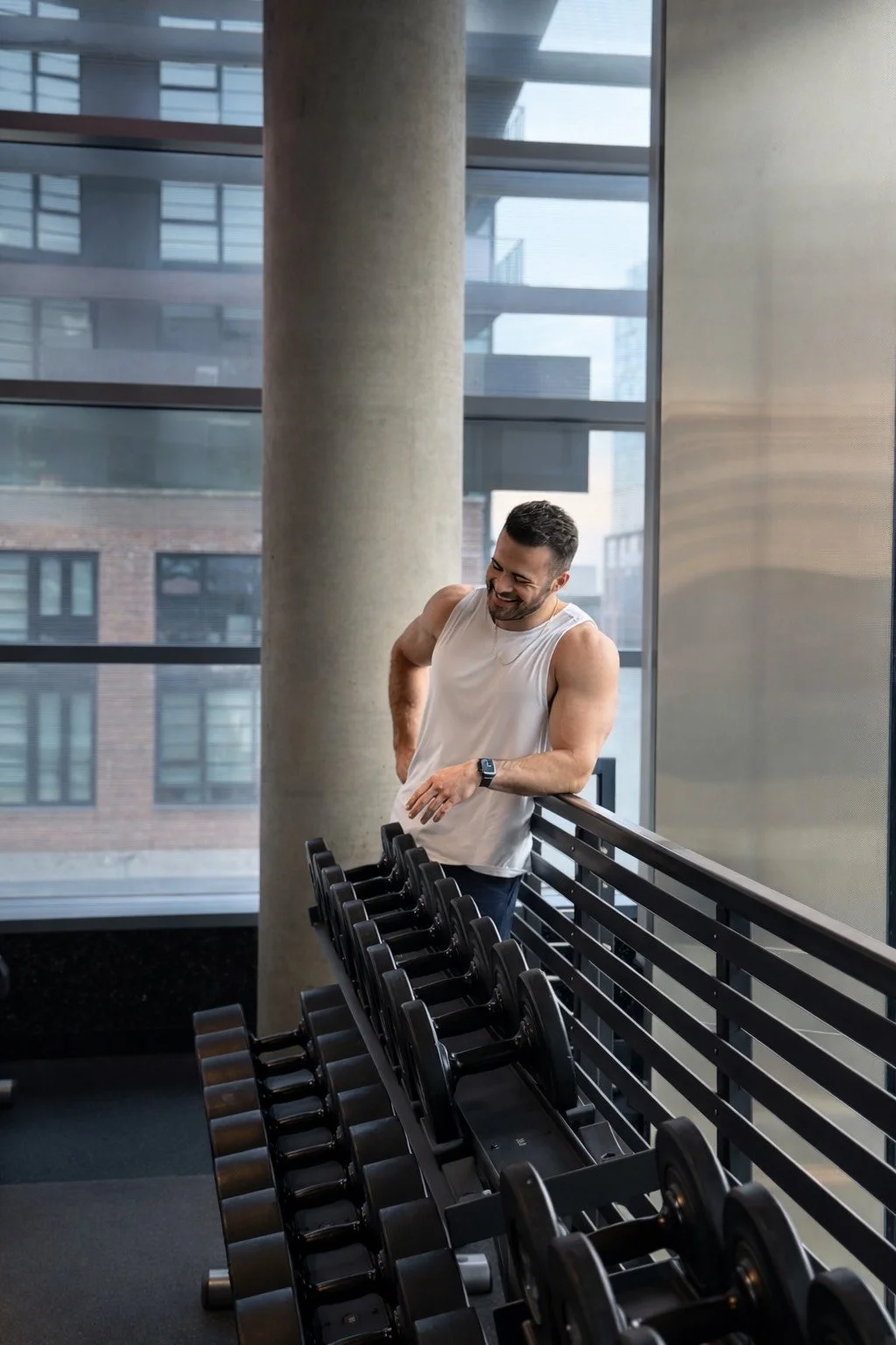 A man in a white sleeveless shirt is smiling and looking down while leaning on a rack of black dumbbells in a gym with large windows.