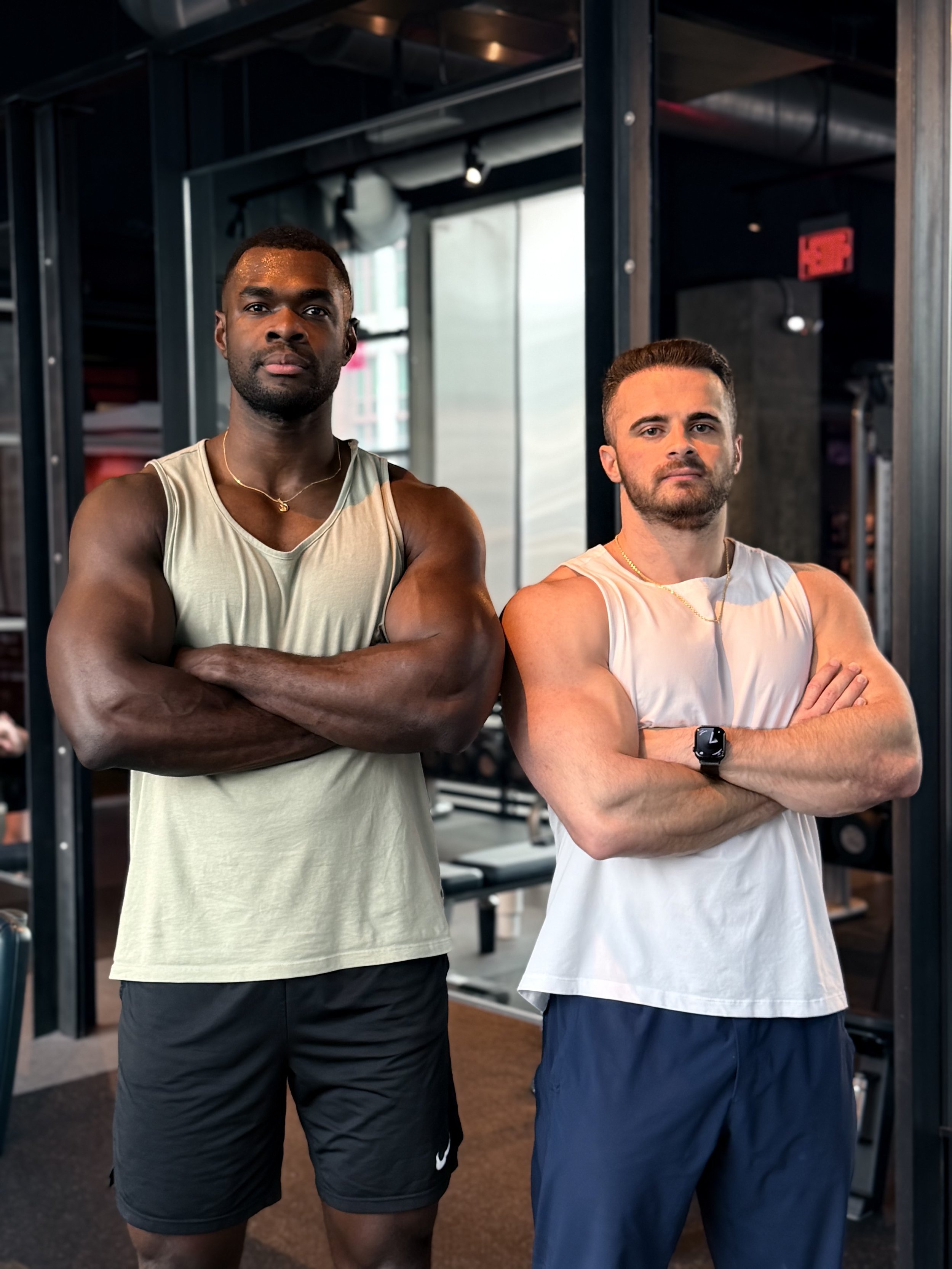 Two muscular men stand with arms crossed in a gym, posed confidently. One is African American with short hair, wearing a beige tank top and black shorts. The other is Caucasian with short hair and a beard, wearing a white tank top, navy pants, and a smartwatch. Gym equipment is visible in the background.