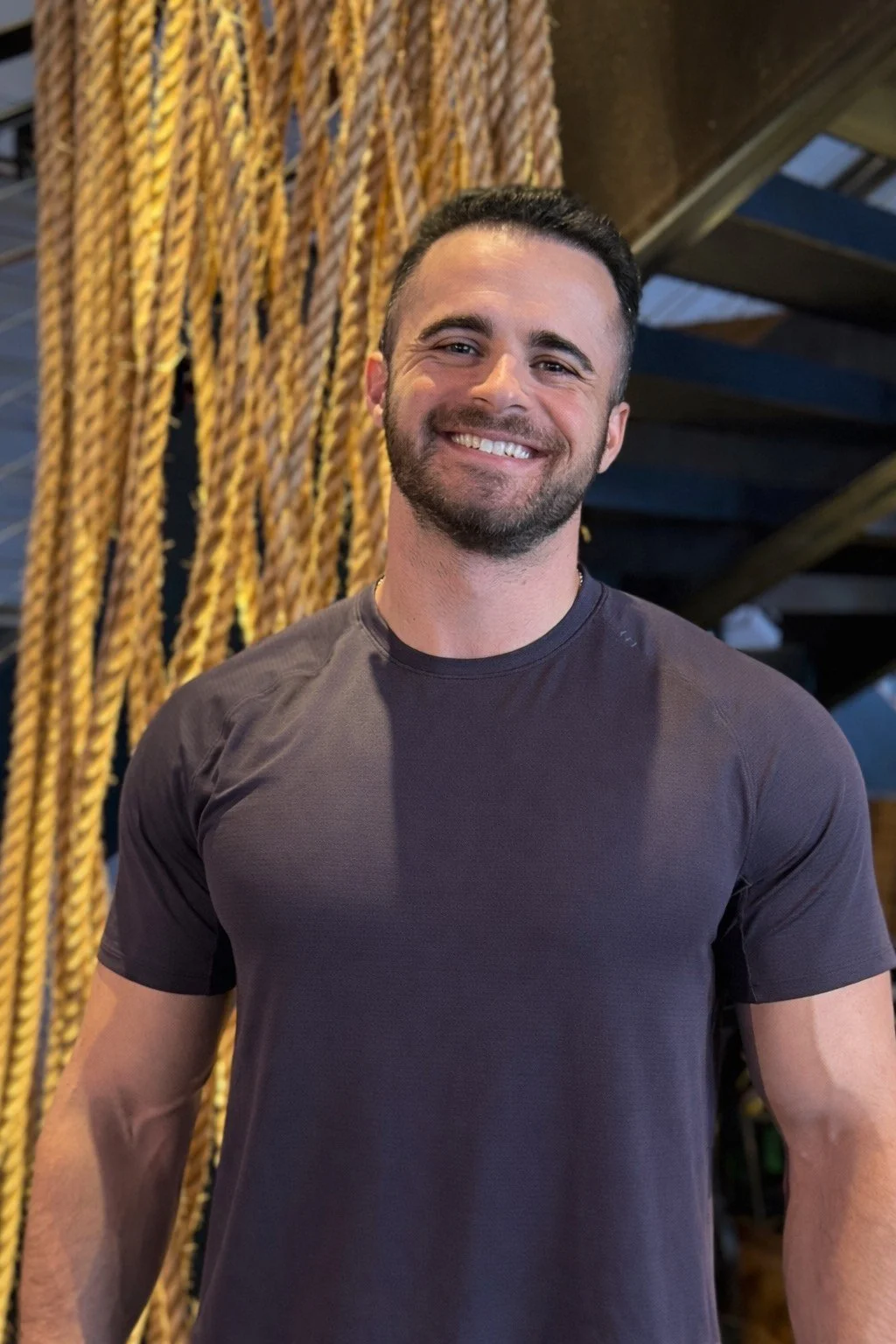 Smiling man with a beard and short dark hair, wearing a dark gray t-shirt, standing in front of a background with yellow ropes or cords hanging vertically.