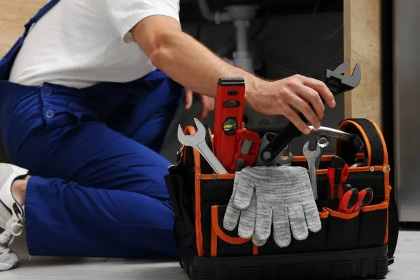 A person kneeling beside a black and orange toolbox filled with various tools including a wrench, a level, a pair of pliers, and a screwdriver, with a pair of gray work gloves hanging over the edge.