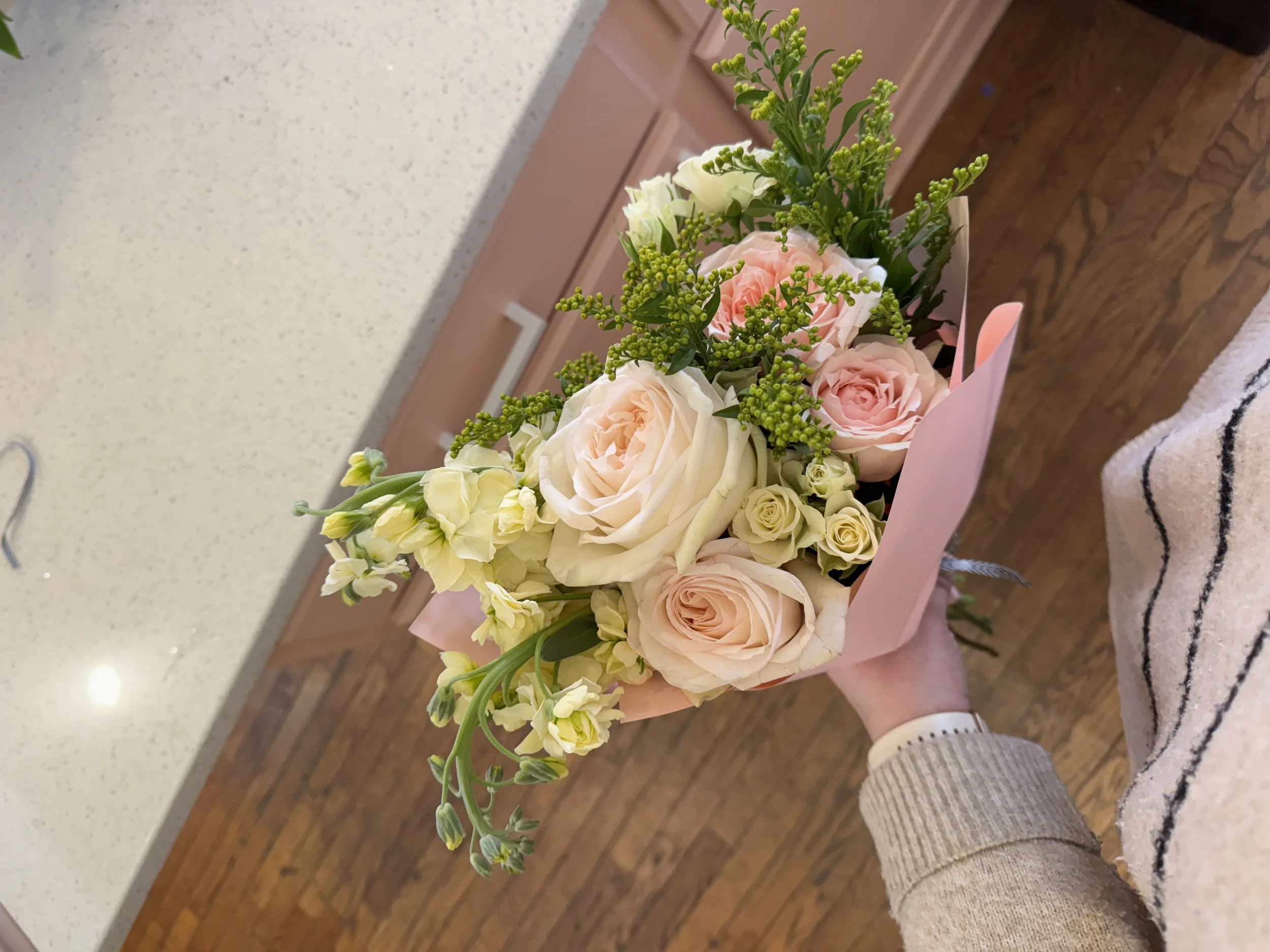 A hand holding a bouquet of pink, white, and cream roses with green foliage and other small flowers, wrapped in pink paper.