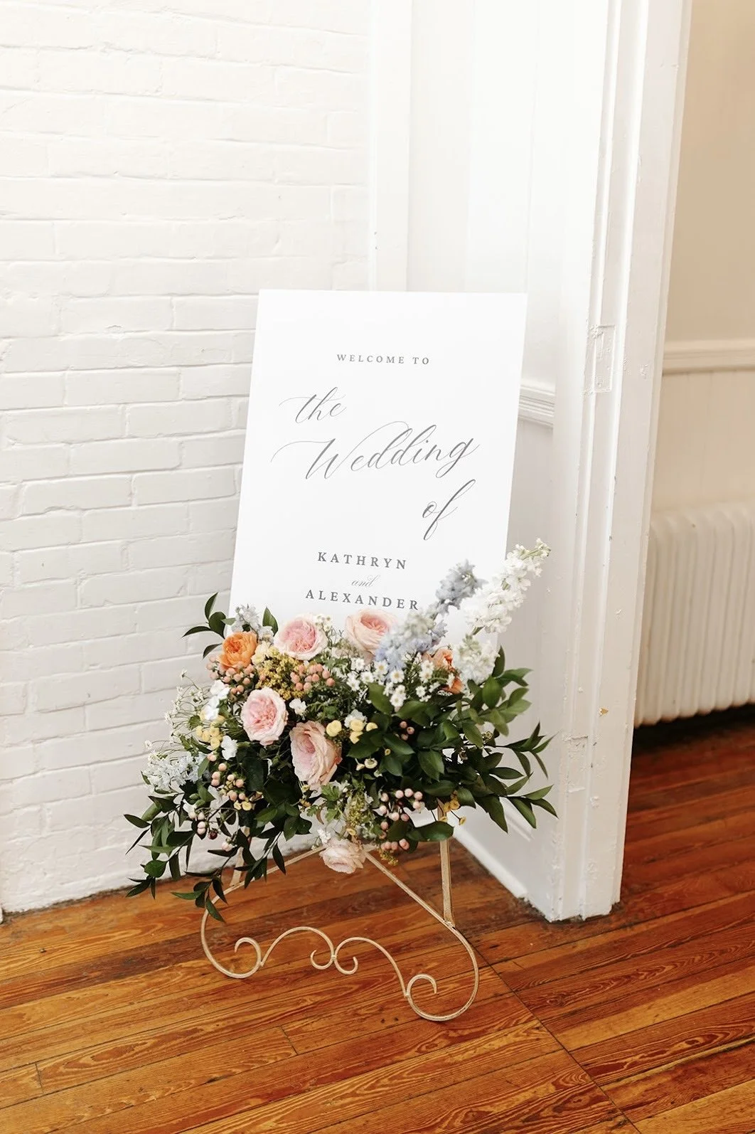 Wedding welcome sign with floral arrangement on a decorative stand in a room with a white brick wall and wooden floor. 1902 venue Orlando Tampa florist