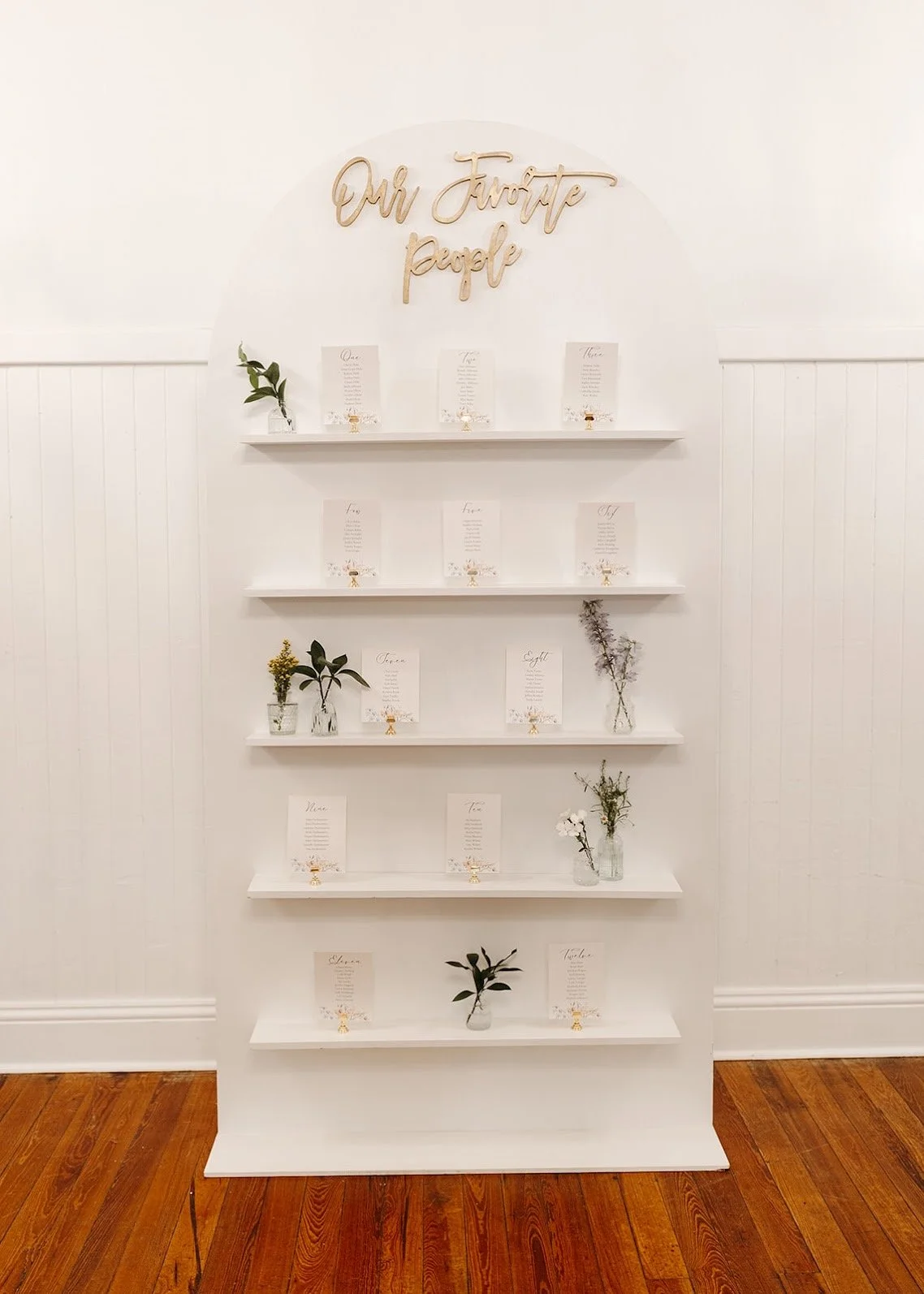 White display shelf with signs and small vases holding greenery, with a gold script sign that reads 'Our Favorite People' at the top.