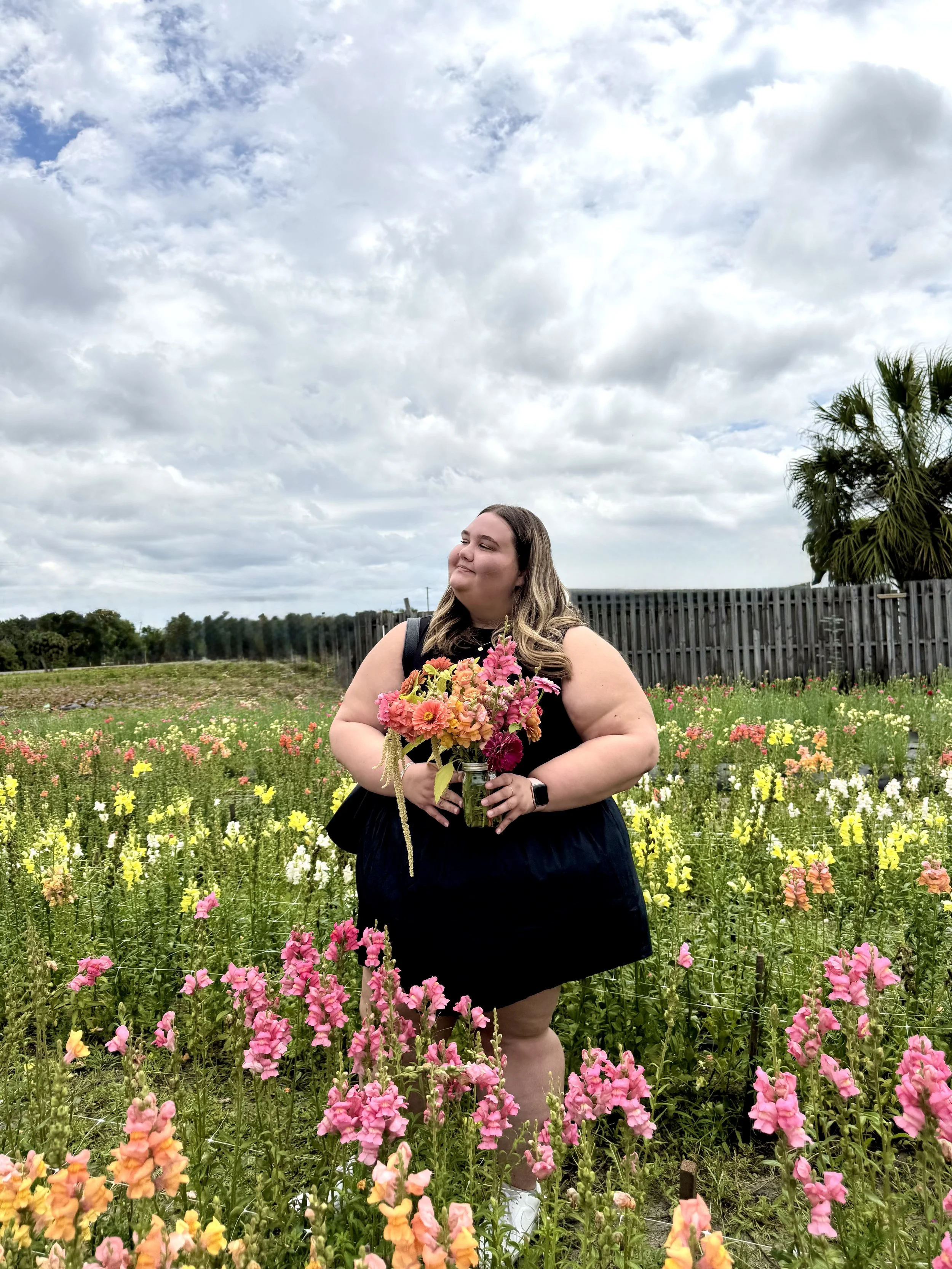 Madison Johnston in a black dress holding a mason jar with pink and orange flowers, standing among a colorful flower field under a cloudy sky. Local Flowers. Lakeland Flowers. Plant City. Tampa. Orlando. Brandon. Event Florist. Wedding Florals.