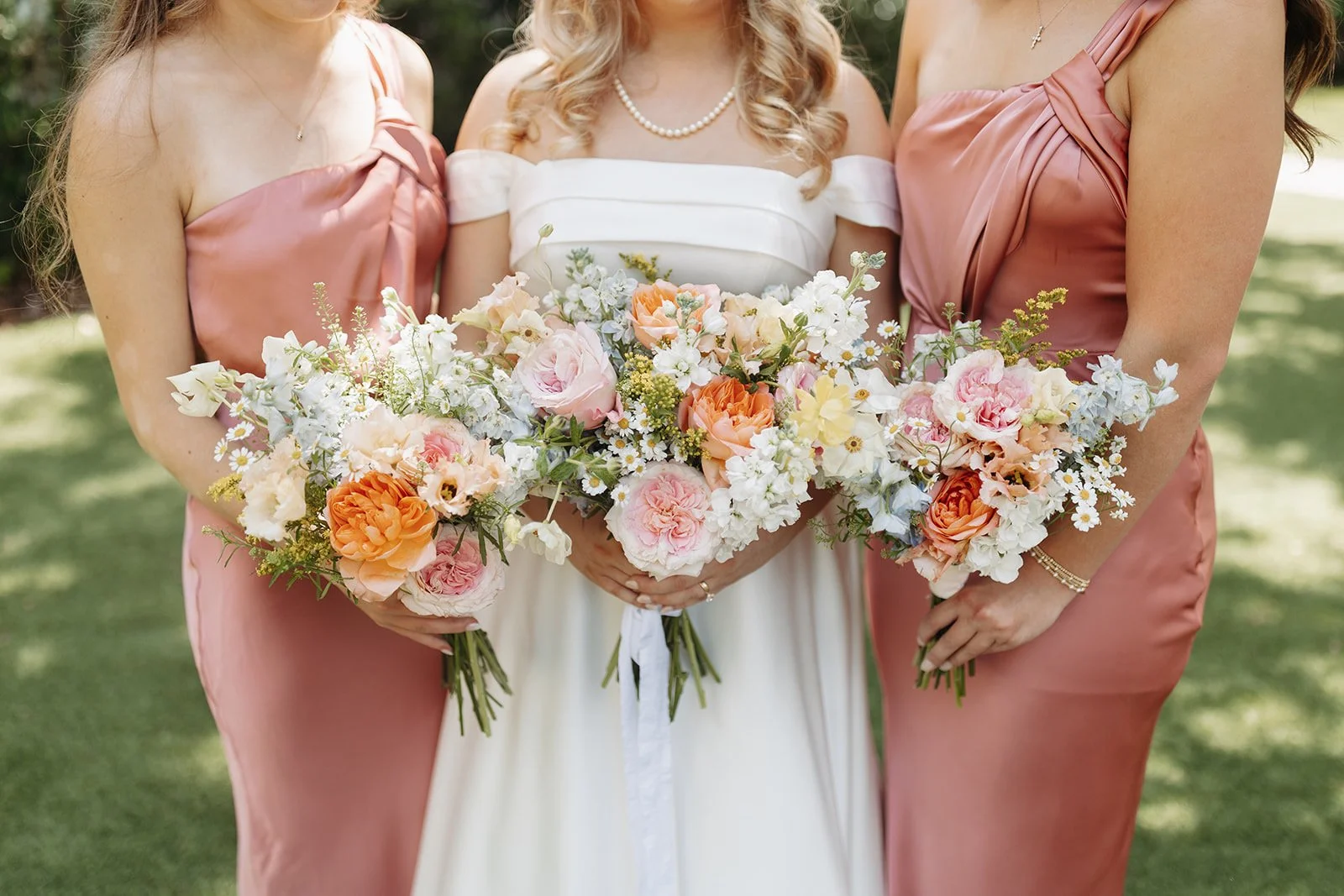 A bride in a white dress holding a bouquet of peach, pink, and white flowers, flanked by two bridesmaids in pink satin dresses holding similar bouquets, outdoors with greenery in the background. Orlando Tampa florist central Florida wedding