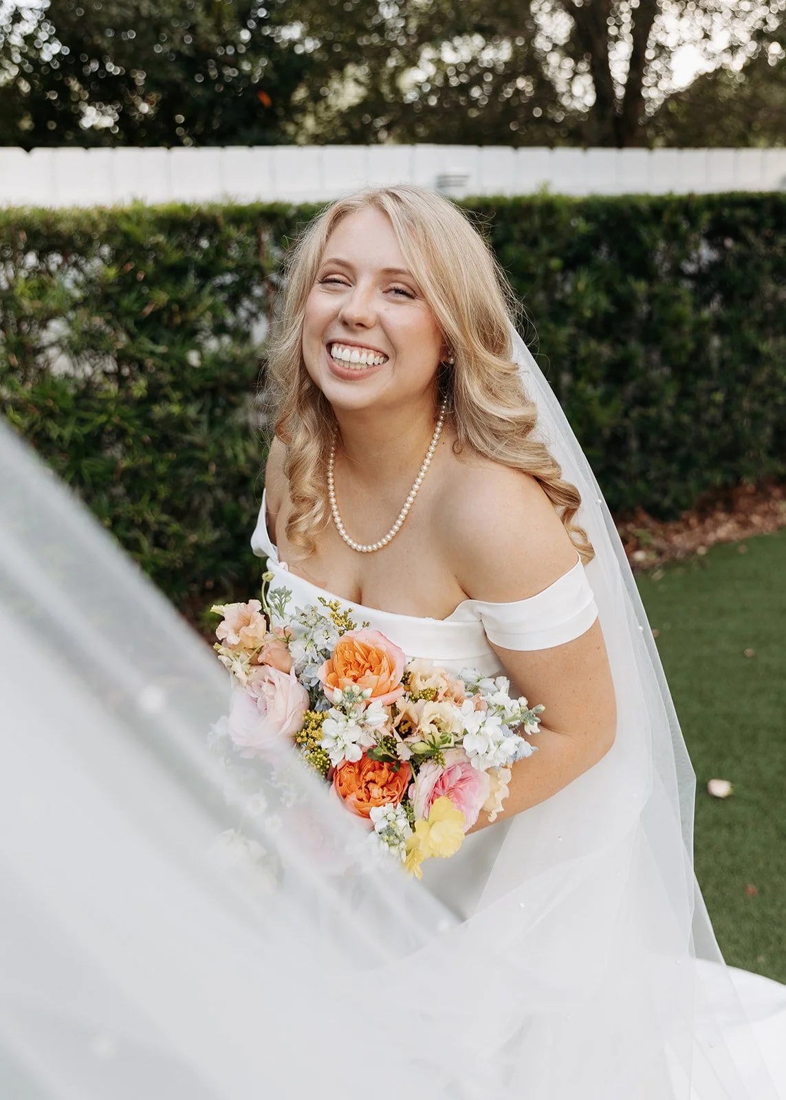 A smiling bride in a white wedding dress holding a bouquet of colorful flowers outdoors.