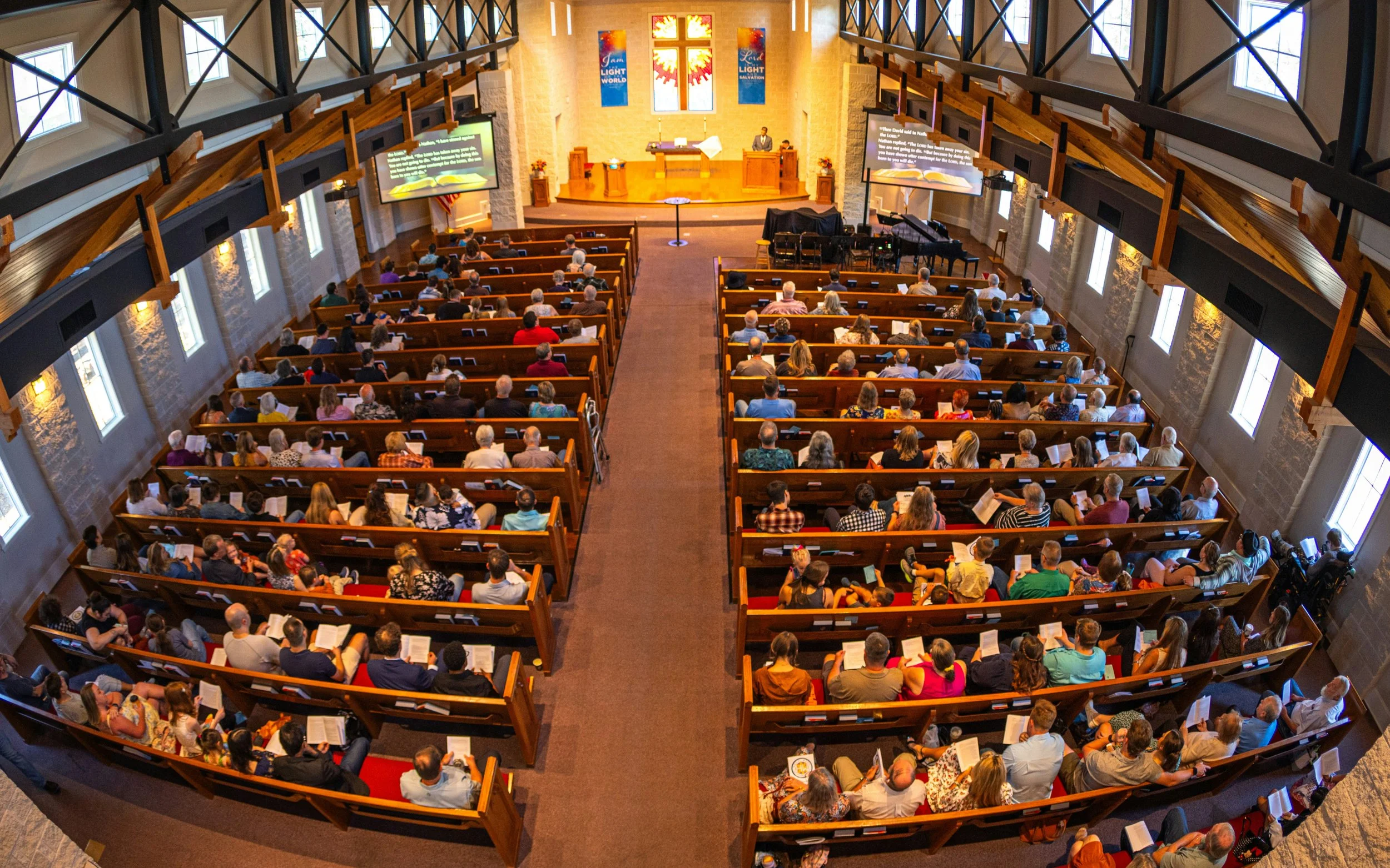 The interior of a church filled with a congregation attending a service, with the altar at the front and stained glass windows behind it.