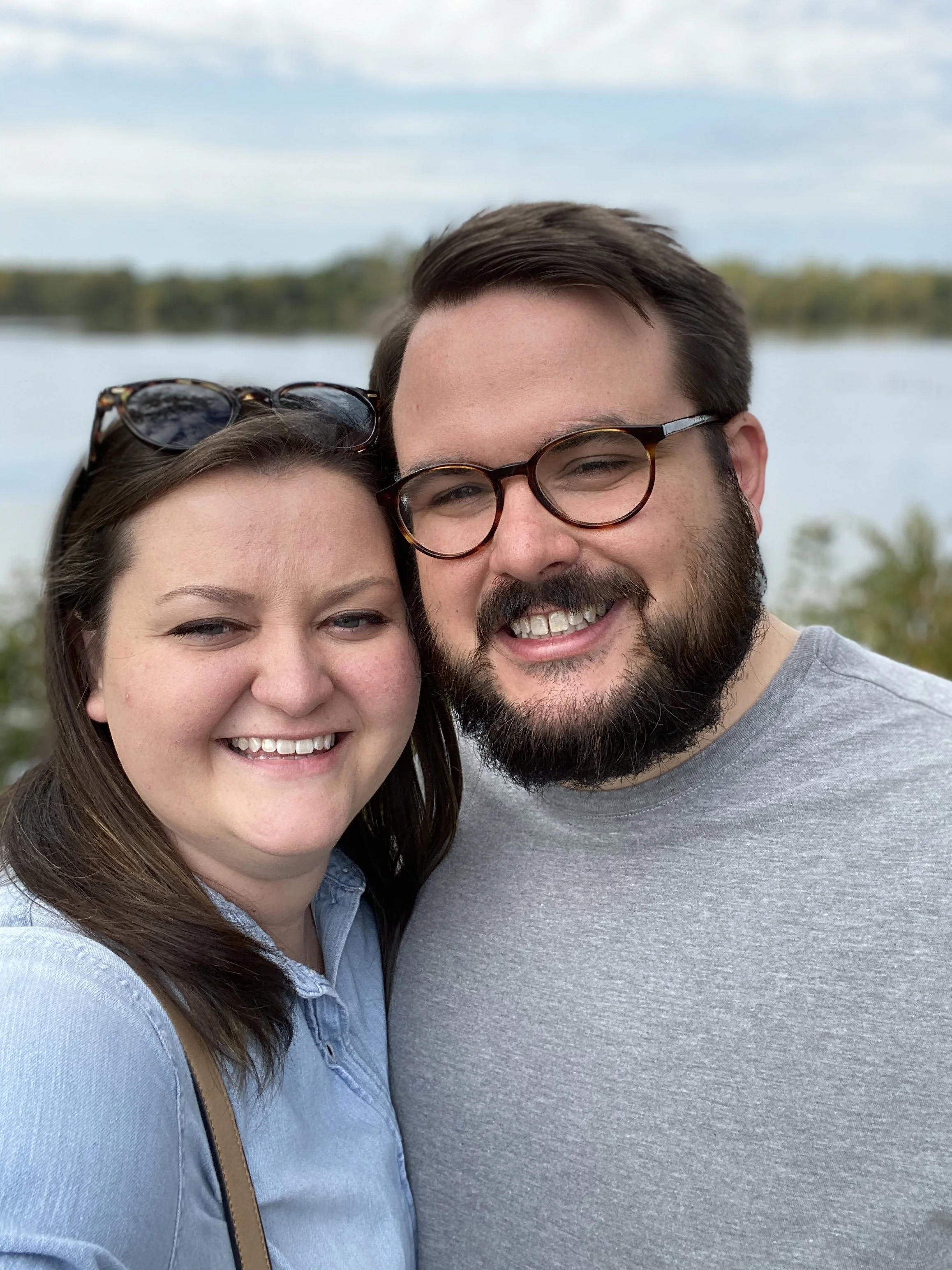 A smiling couple taking a selfie outdoors near a body of water, with trees and a cloudy sky in the background.