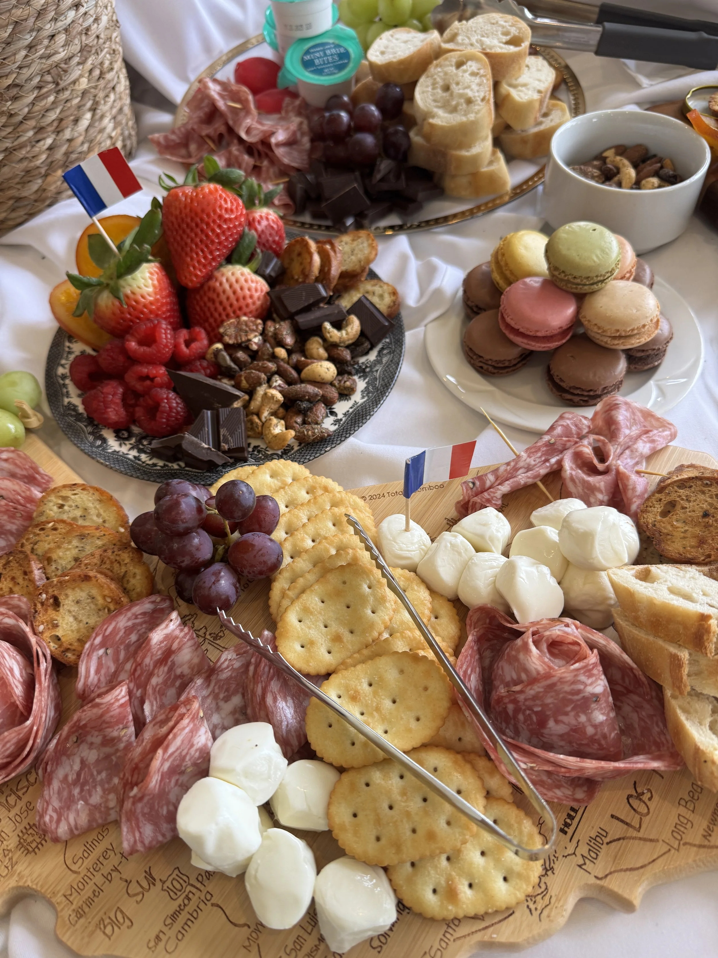 A charcuterie board with various cured meats, cheese, crackers, grapes, and other finger foods, along with a platter of fresh fruits, chocolates, macarons, and a bowl of nuts, all arranged on a white tablecloth for a buffet-style meal.