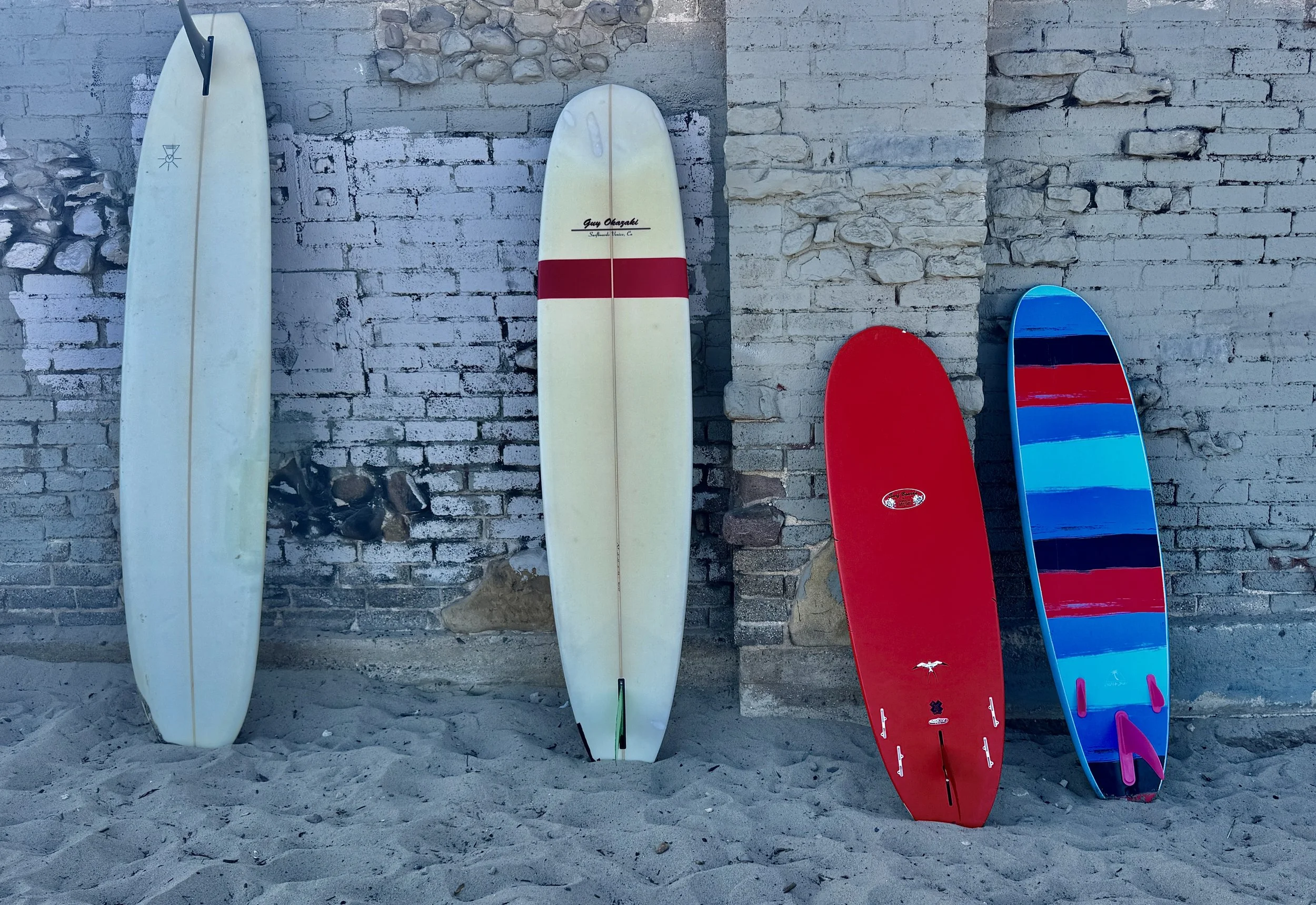 Four surfboards leaning against a gray brick wall on sandy ground.