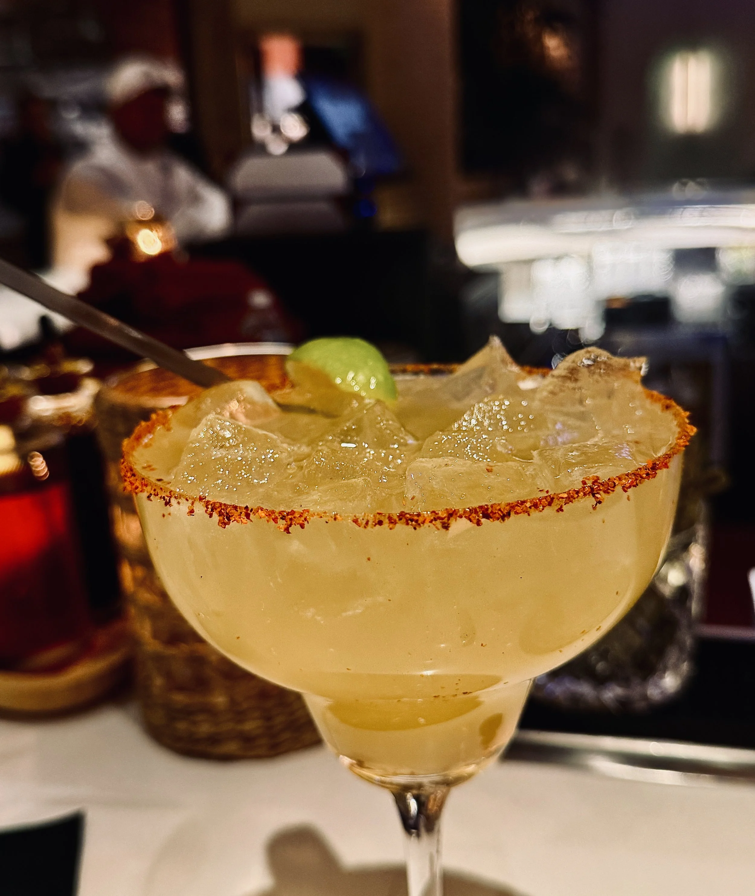 A margarita cocktail in a glass with a salted rim and lime garnish, filled with ice and a pale yellow liquid.
