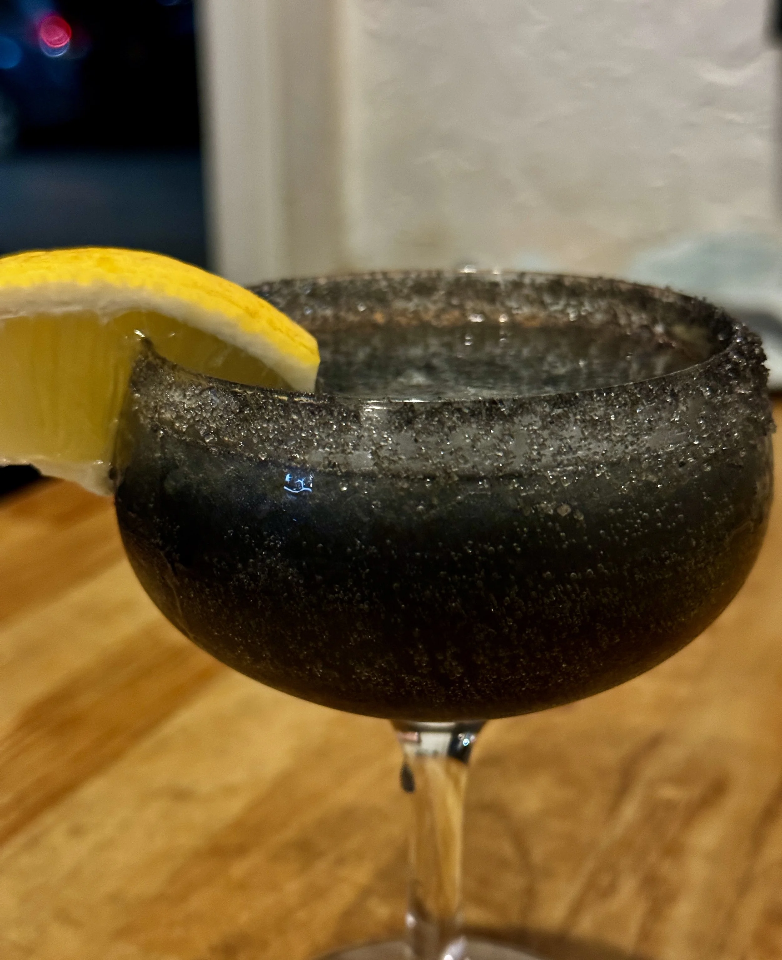 A close-up of a black cocktail glass with a salted rim and a lemon wedge garnish.