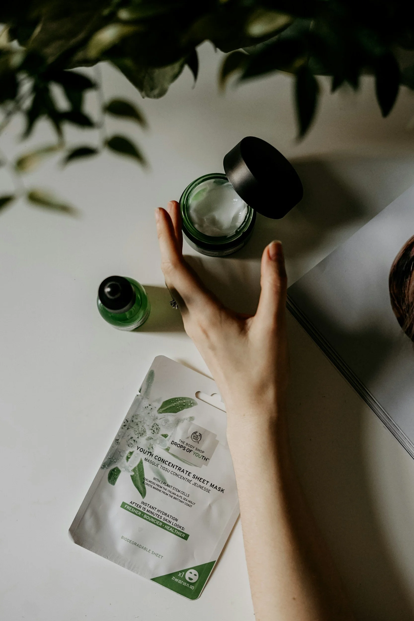 A person holding a jar of skincare balm or cream, with an open face mask packet on a white surface, a small green bottle nearby, and a plant in the background.