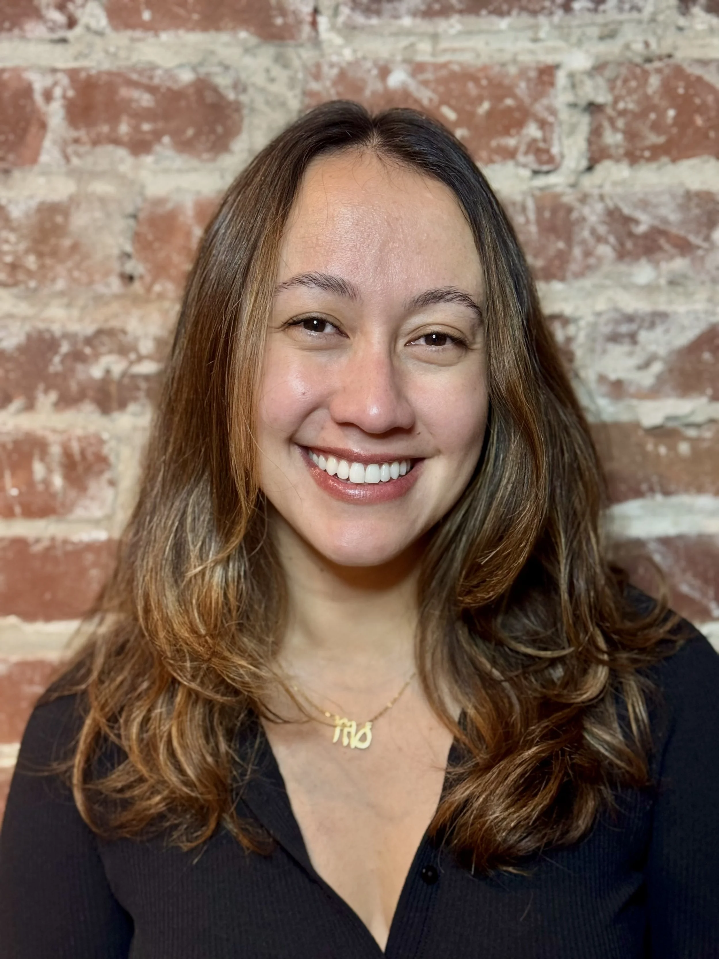 A smiling woman with long, wavy brown hair wearing a black top and a gold necklace with the initials 'TMD' in front of a brick wall.