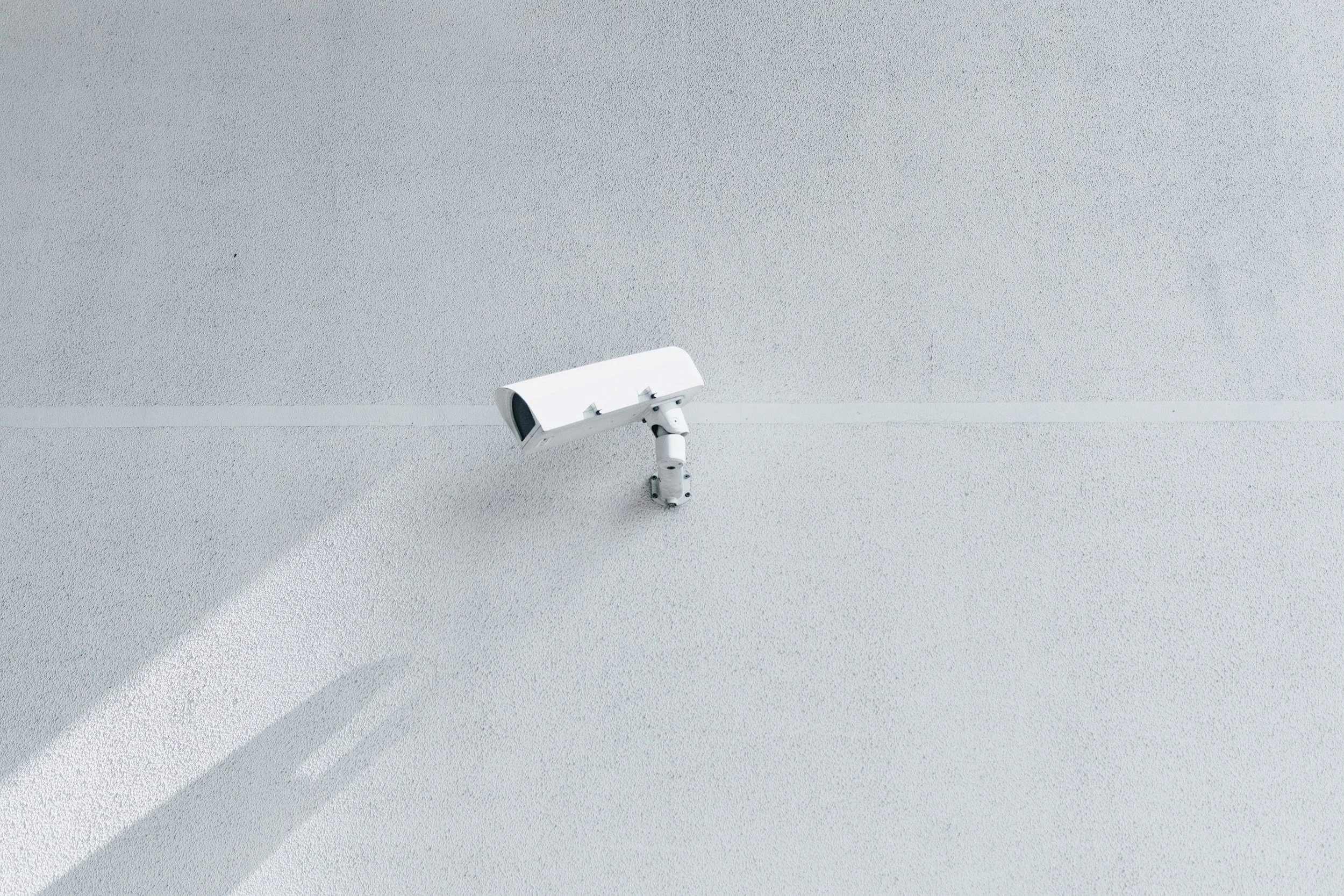 A white security camera mounted on a plain white wall, casting a shadow.