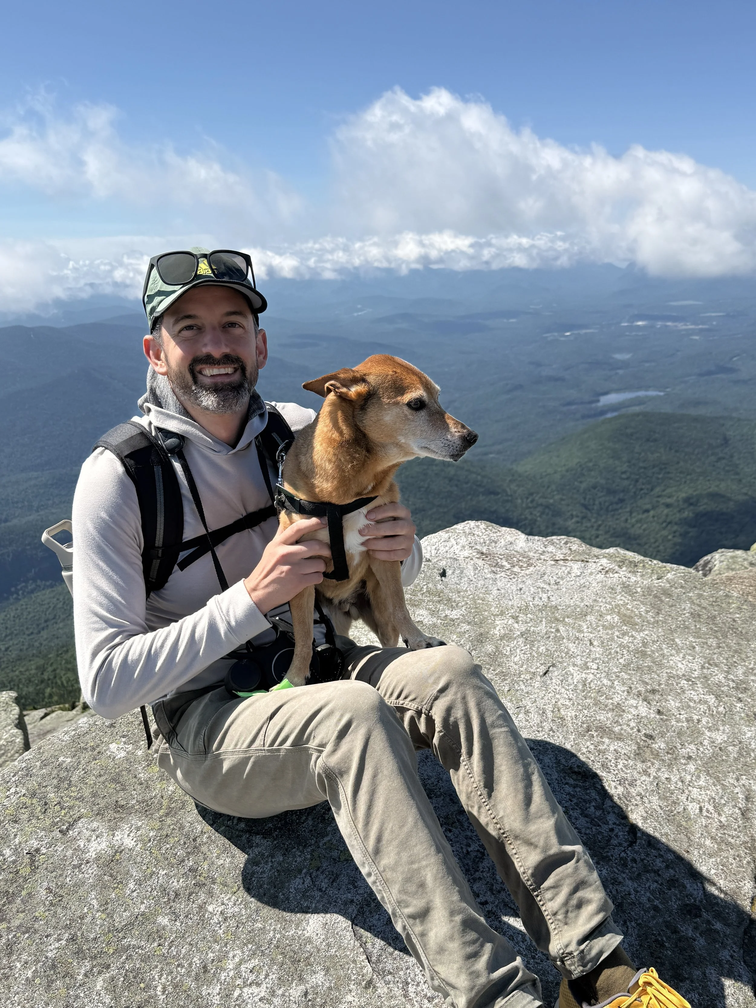 A man with a beard, wearing a hat with sunglasses on top, and a backpack, sitting on a rock on a mountain summit, holding a small brown dog while overlooking a landscape of mountains, forests, and clouds in the distance.