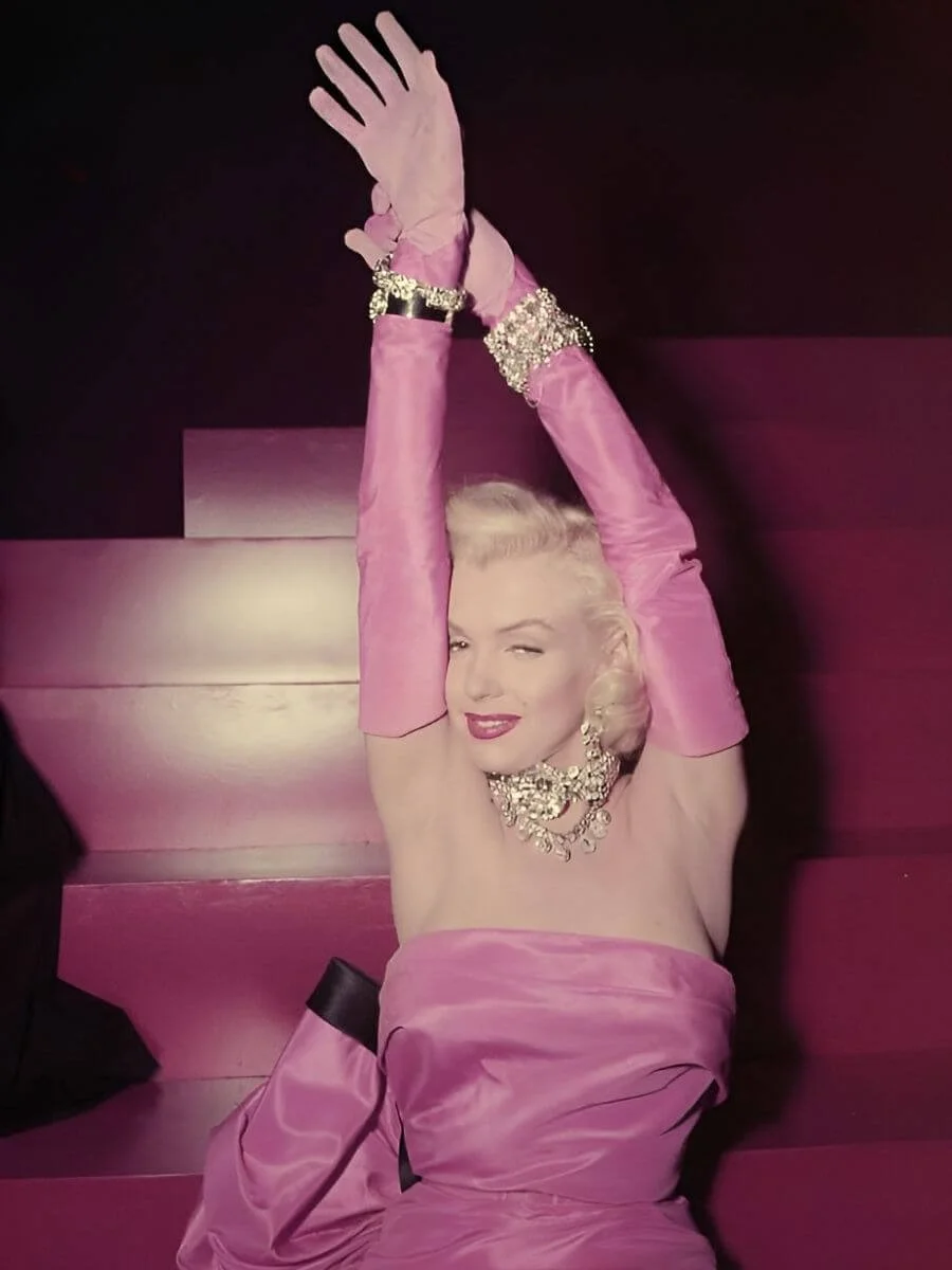 Marilyn Monroe in a pink gown with matching gloves against a red backdrop