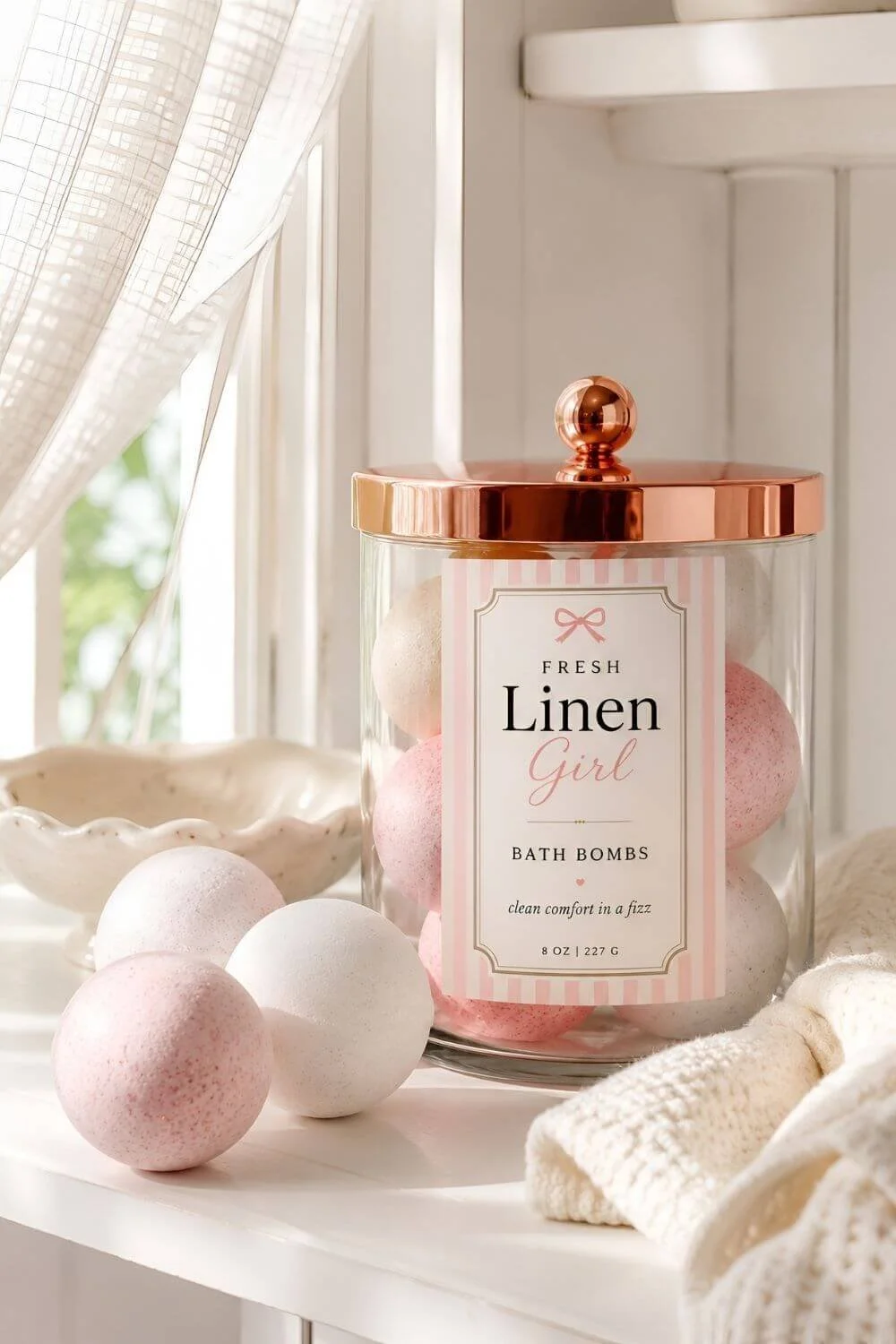 Fresh Linen Girl bath bombs in a clear glass jar with a rose gold lid, styled on a white shelf with soft knit fabric, loose pink and white bath bombs, and natural window light.