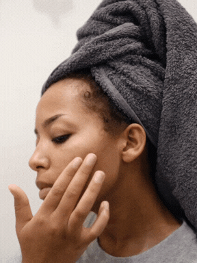 A woman with a towel wrapped around her head touching her face, possibly in a skincare routine.