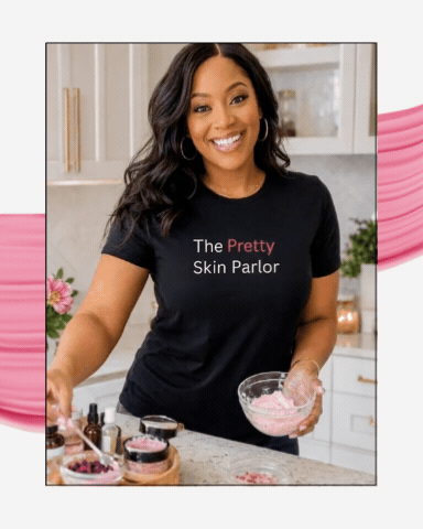 Woman smiling in a kitchen holding a bowl of pink bath salts or skincare product, with skincare jars and flowers on the counter, wearing a black t-shirt that says