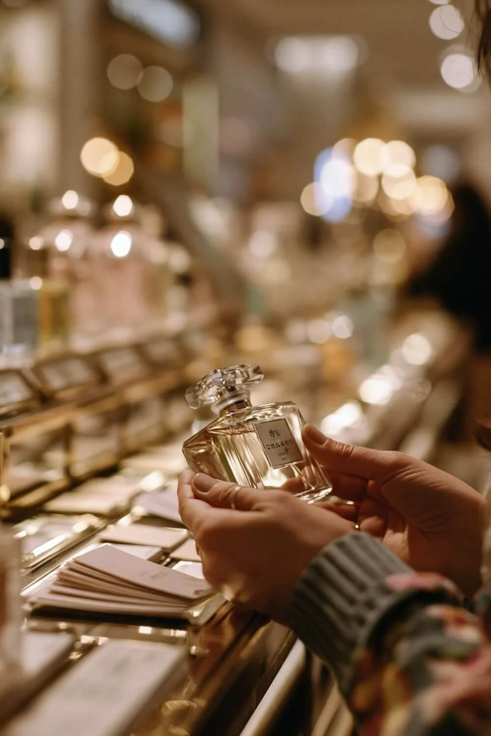 Woman holding a luxury perfume bottle in a softly lit fragrance boutique with warm golden bokeh lights.