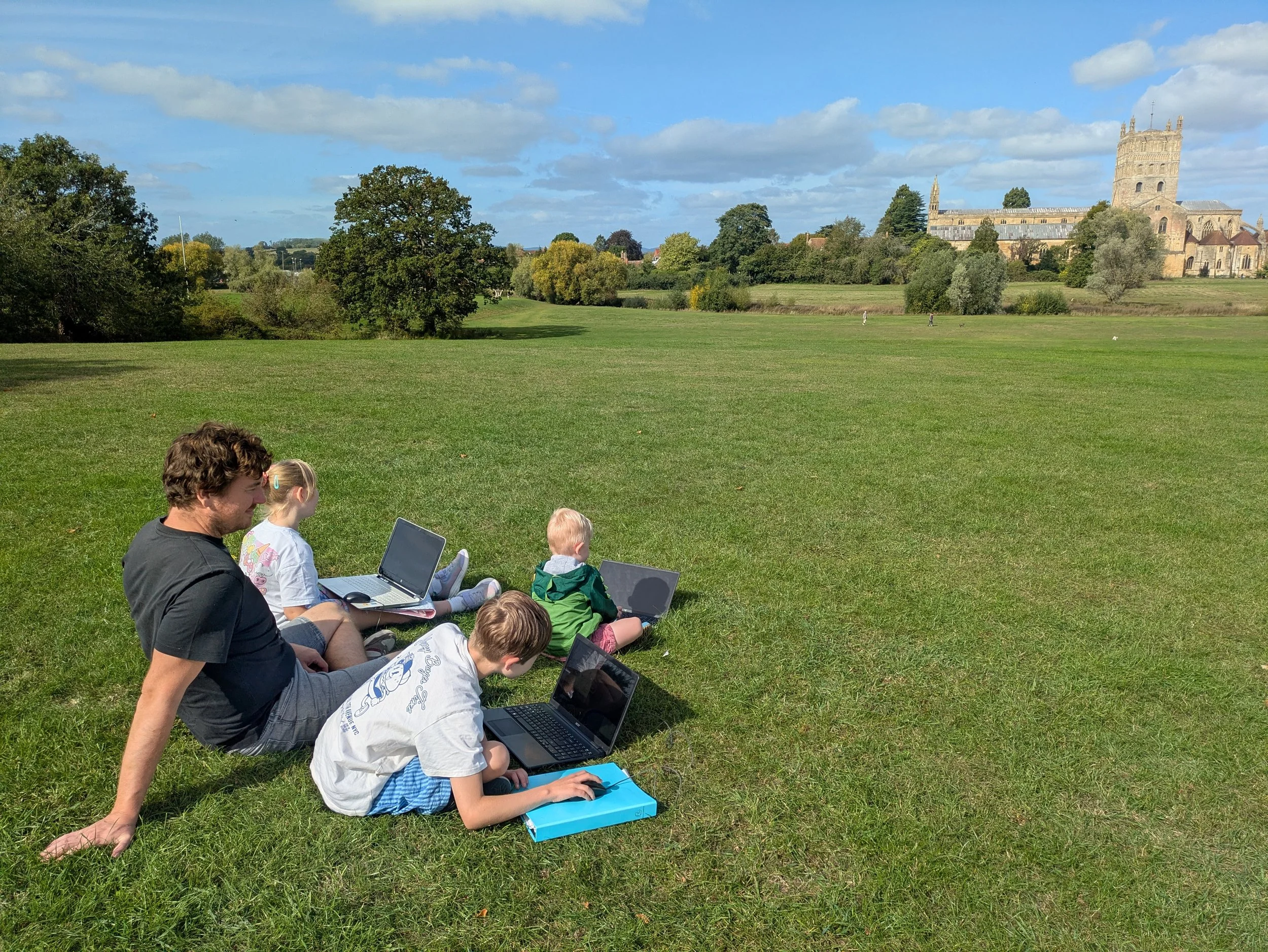 Several children and a man sitting on grass with laptops outdoors in front of historic buildings and trees on a bright, partly cloudy day.