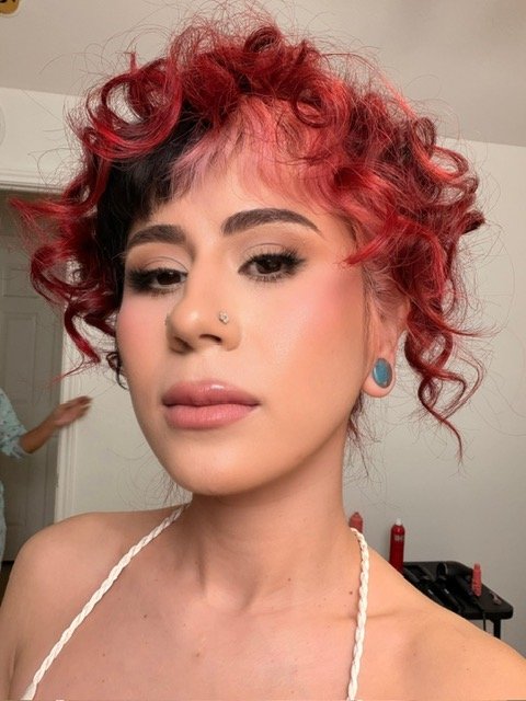 A woman with short, curly hair styled in a mix of black and vibrant red. She has makeup with natural lips, winged eyeliner, and well-defined eyebrows. She has a nose piercing and blue ear gauges. She is wearing a white rope strap top, and the background shows a plain wall and a person partially visible.