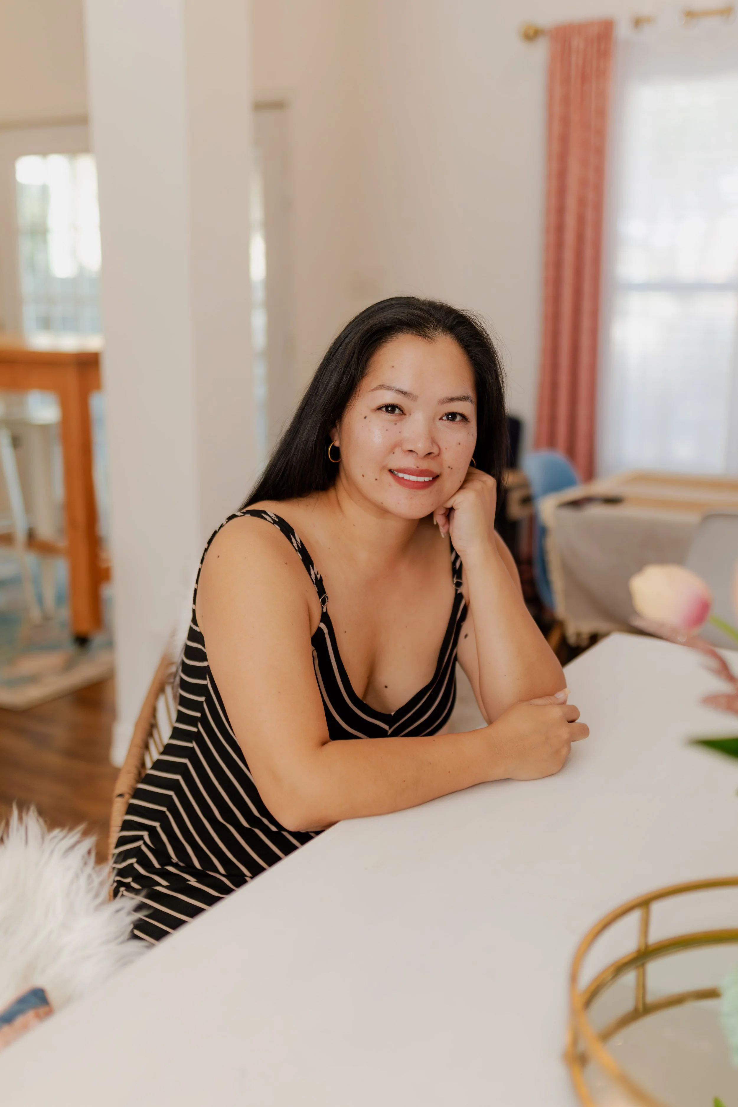 A woman with long black hair wearing a black and white striped sleeveless dress sits at a table, smiling at the camera in a well-lit room with pink curtains and a window in the background.