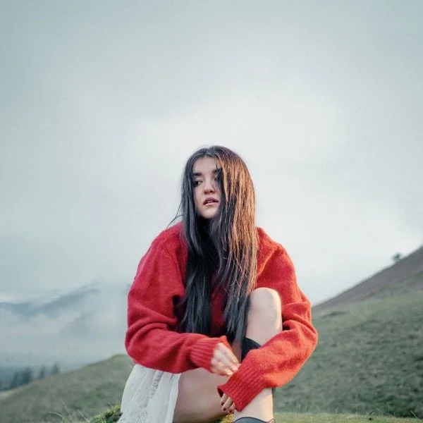 A young woman with long dark hair sits outdoors on grass in a landscape with hills and misty mountains in the background, wearing a red sweater.