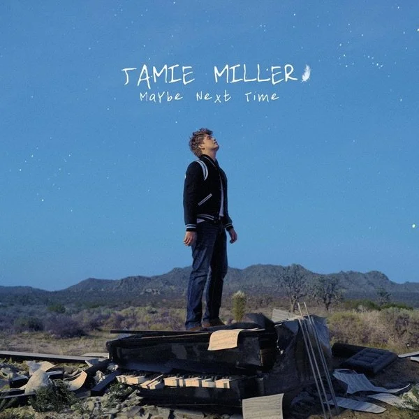 A young boy stands on a toppled piano in a desert landscape under a starry night sky, looking up at the sky. Handwritten text above him reads "JAMIE MILLER Maybe Next Time."