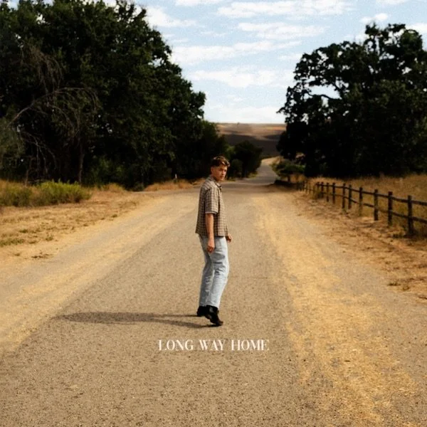 A young man in a plaid shirt and jeans walking on a dirt road with trees and fences on either side, under a partly cloudy sky, with the words "LONG WAY HOME" at the bottom.