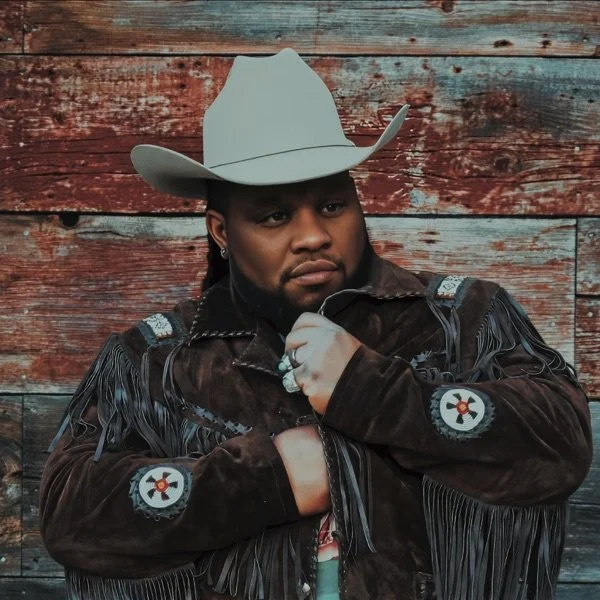 Man wearing a cowboy hat and fringe leather jacket, standing in front of a wooden wall