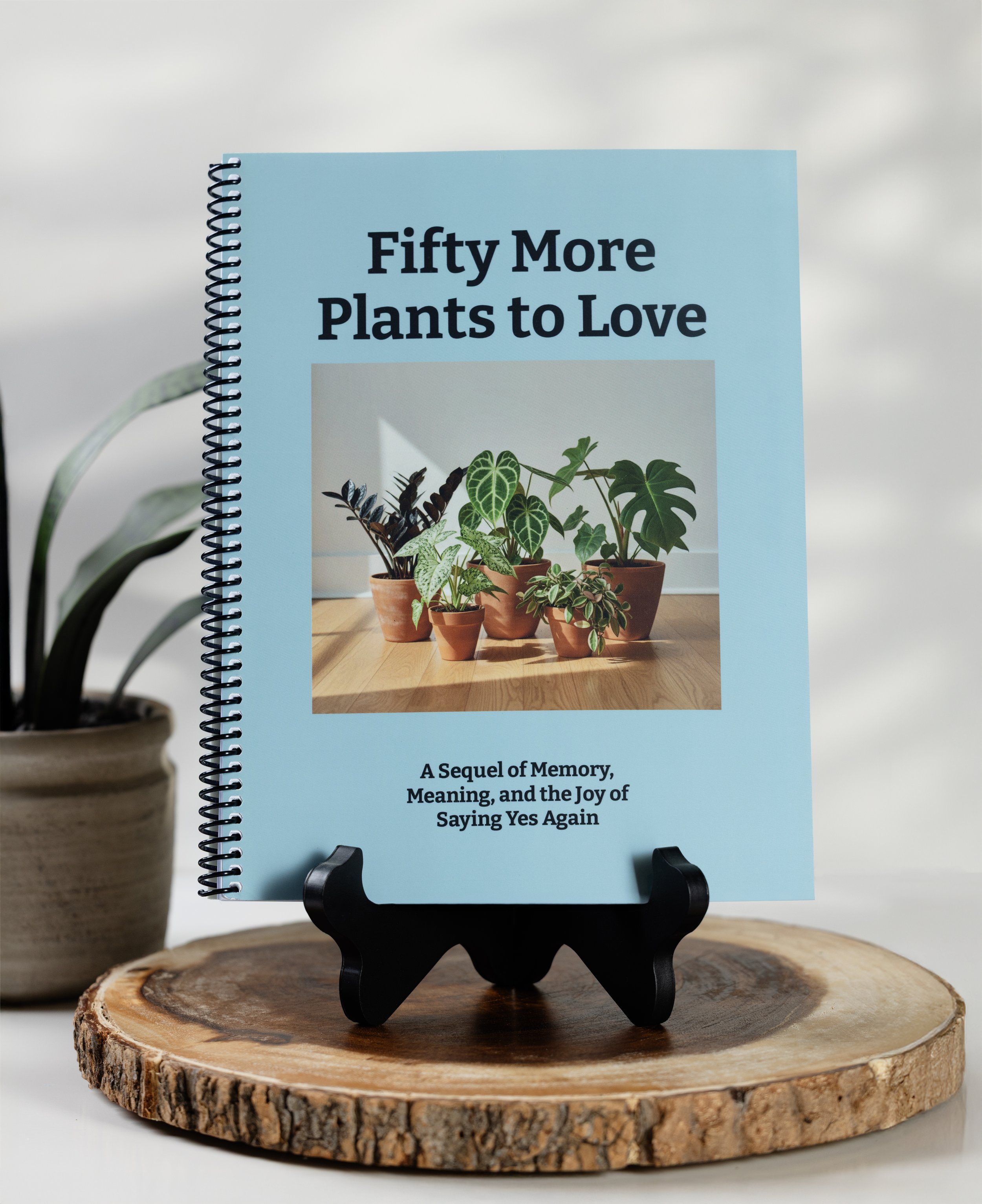 A book titled "Fifty More Plants to Love" displayed on a black book stand on a wooden surface, with a potted plant partially visible on the left and a light-colored background.