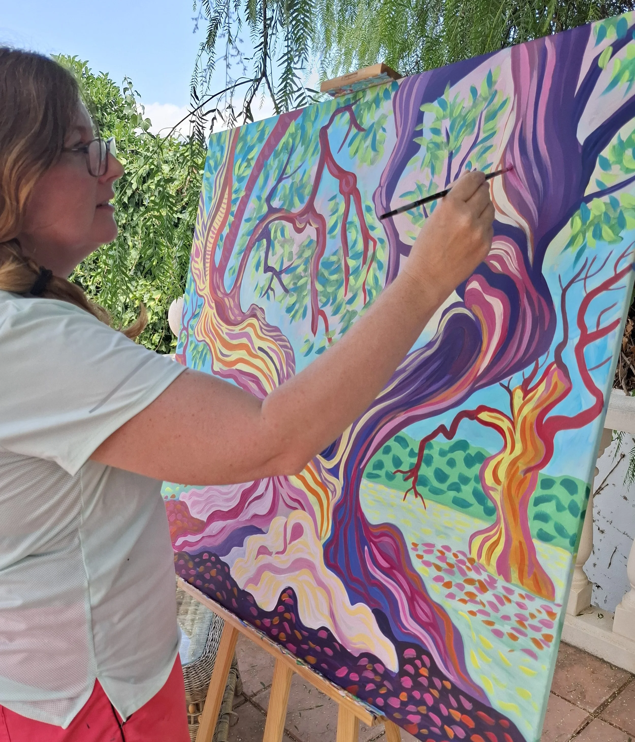 A woman painting a colorful, abstract landscape of trees and foliage on a large canvas outdoors.