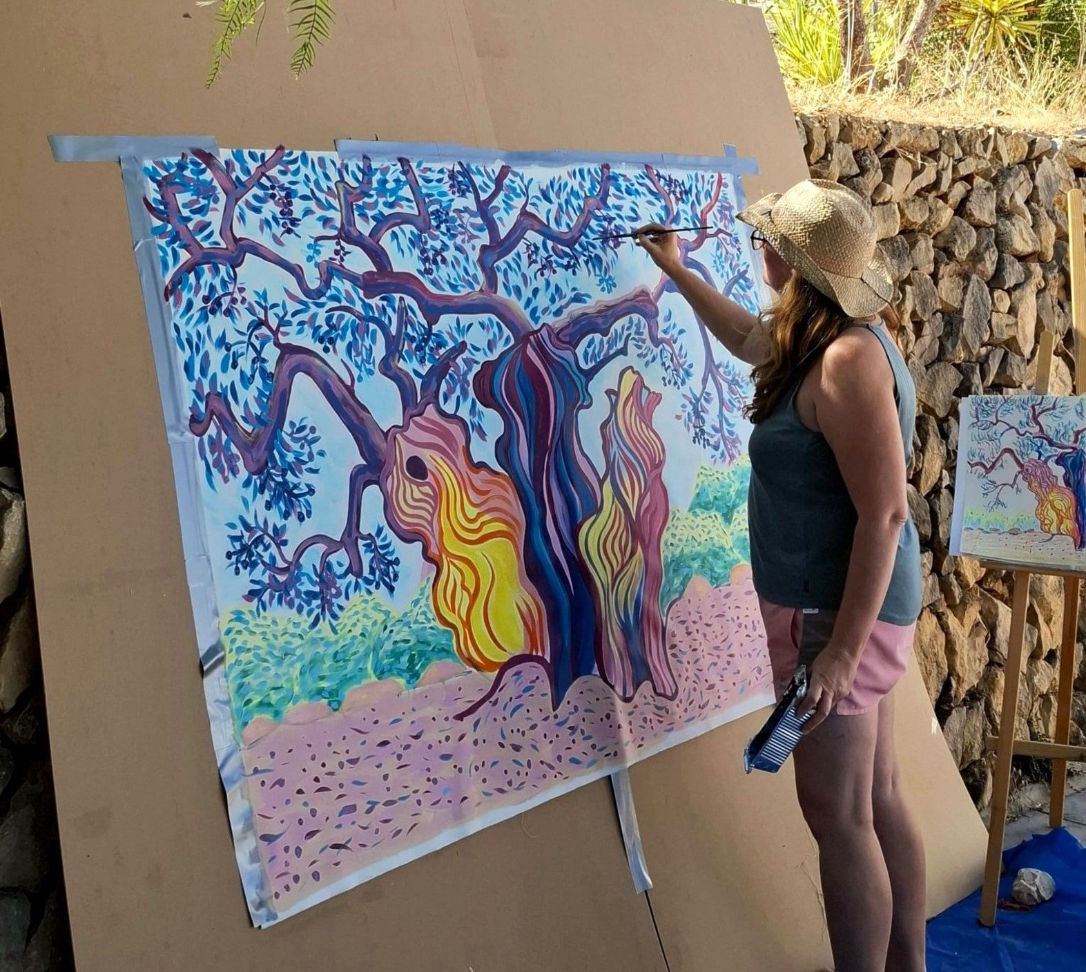 A woman with long brown hair wearing a straw hat, a dark sleeveless top, and pink shorts is painting a colorful abstract tree on a large canvas outdoors. She is holding a paintbrush in one hand and a palette or paint tray in the other.