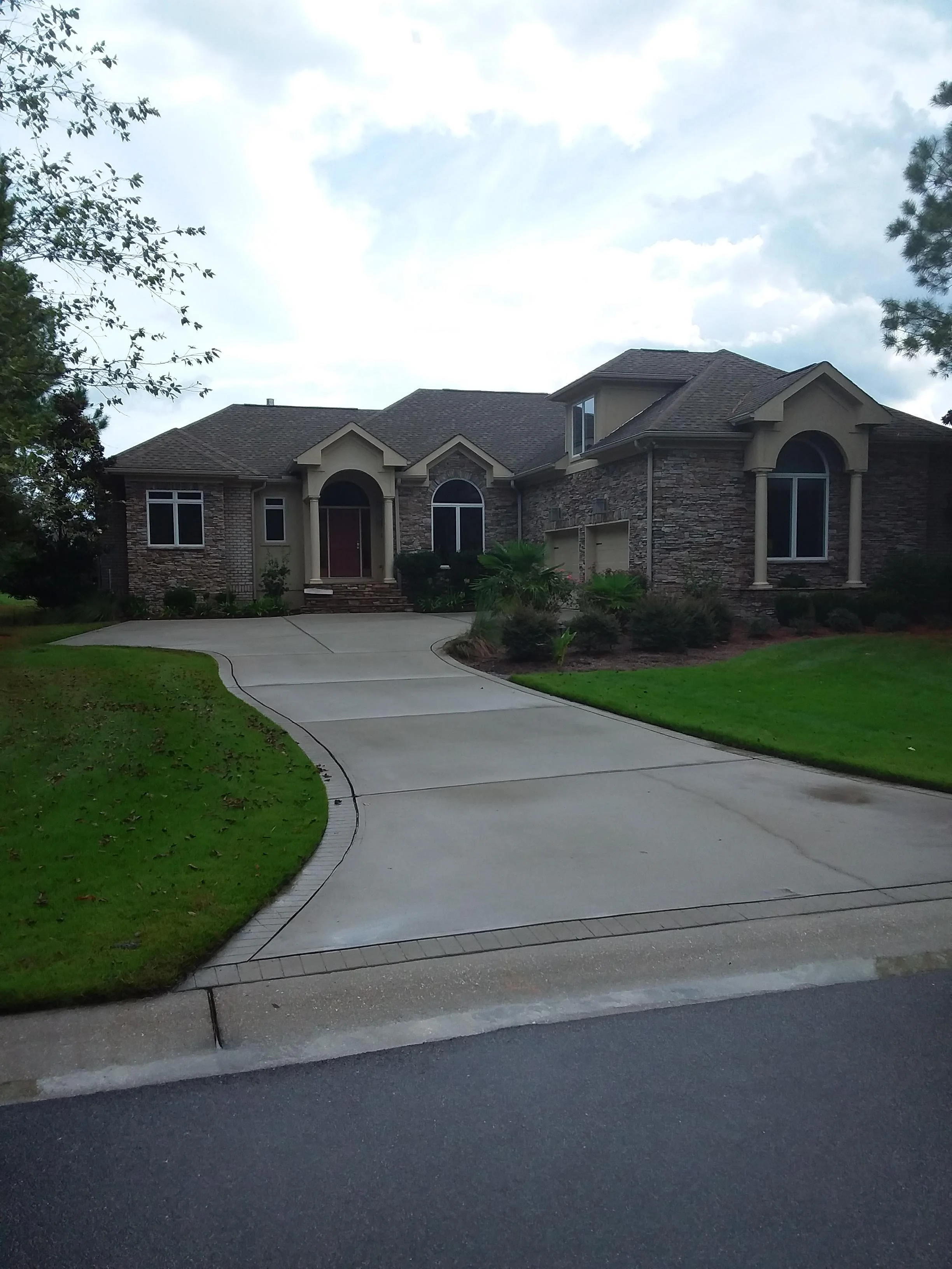 A suburban house with stone and stucco exterior, arched windows, and a two-car garage. A curved driveway leads to the house, surrounded by a well-manicured lawn and landscaping. The sky is partly cloudy.