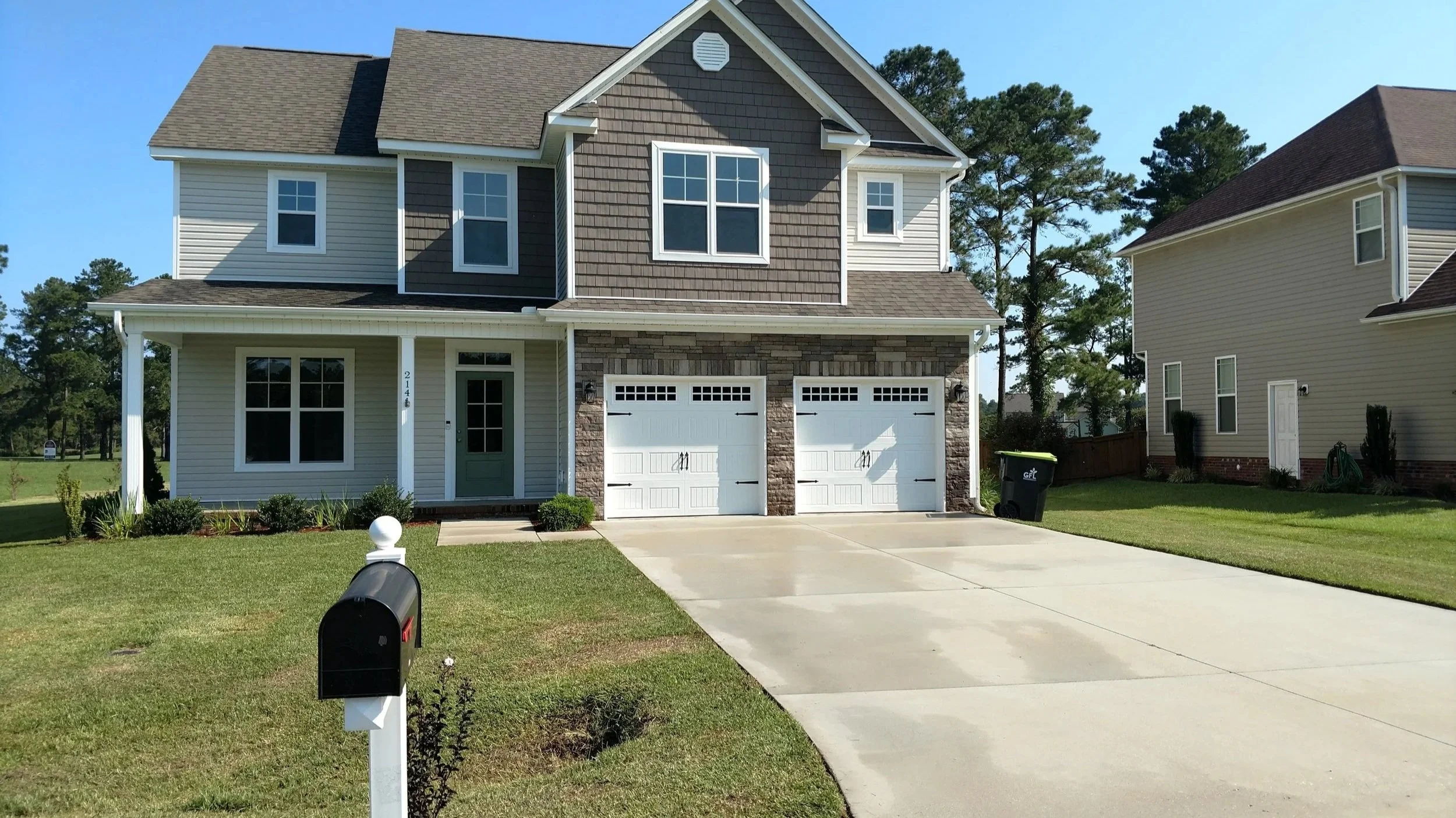 A two-story suburban house with beige and brown exterior. It has a two-car garage, a green front door, and multiple windows. The front yard has a manicured lawn, small shrubs, and a concrete driveway. A mailbox is in the foreground, and neighboring houses are visible in the background. Tall trees are behind the house.