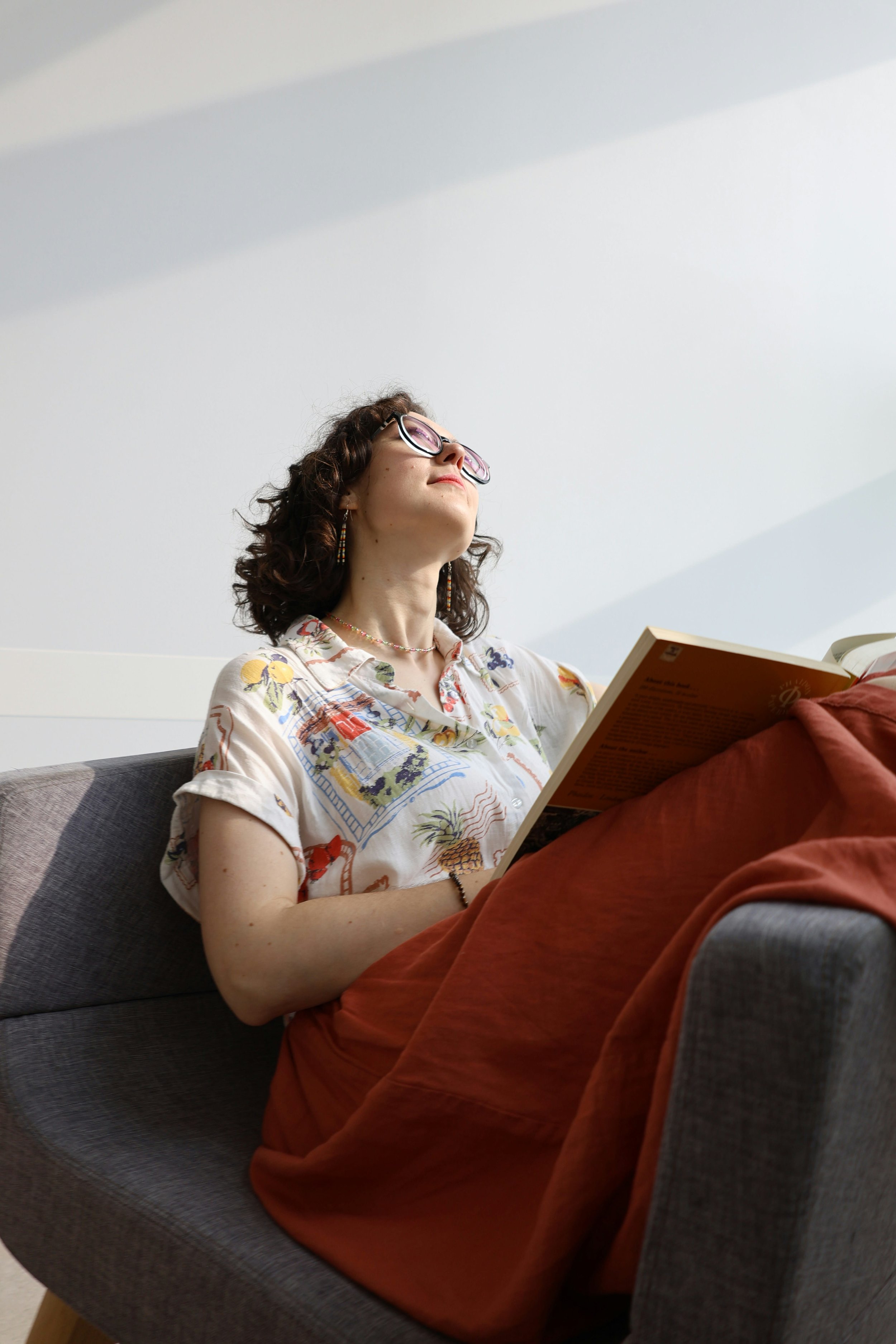 A woman with curly dark hair wearing glasses and a patterned shirt, sitting on a gray couch, reading a book with a wooden cover. She has a colorful necklace, earrings, and a bracelet.