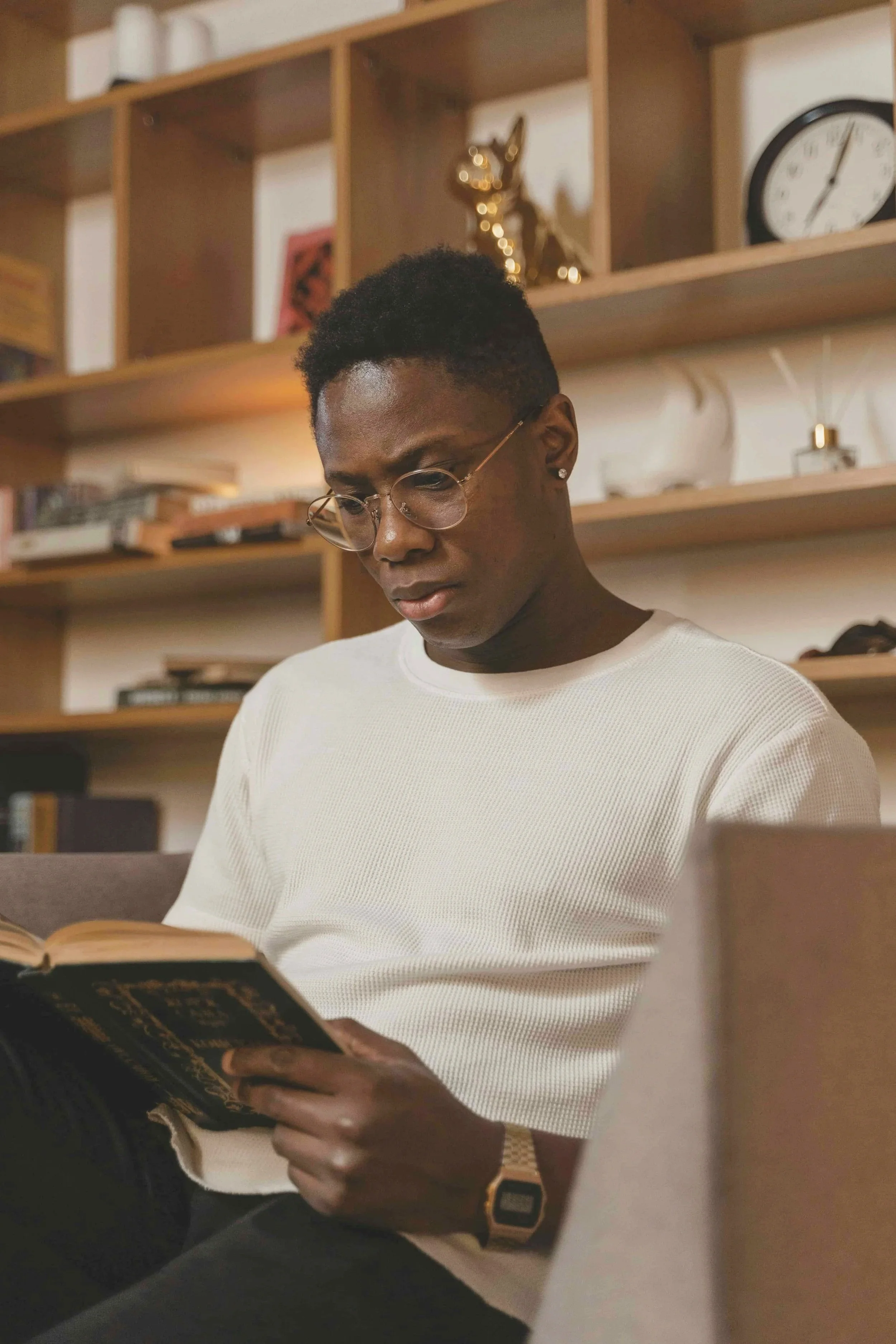 A young man with glasses and a white shirt reading a book in a room with wooden shelves, decorative items, and a clock on the wall.