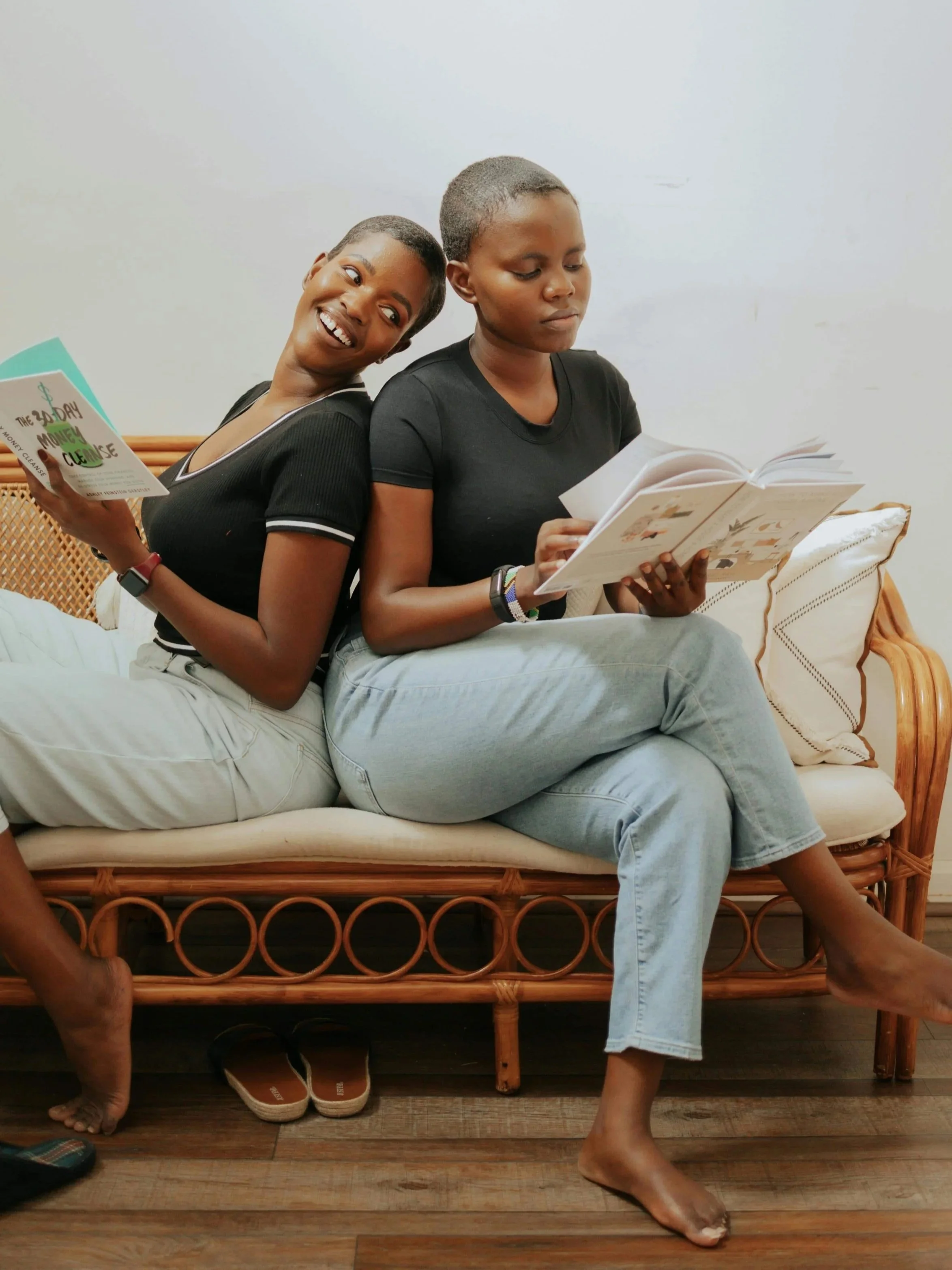 Two women are sitting on a rattan sofa, back-to-back, reading books, with one smiling and the other focused on her book.
