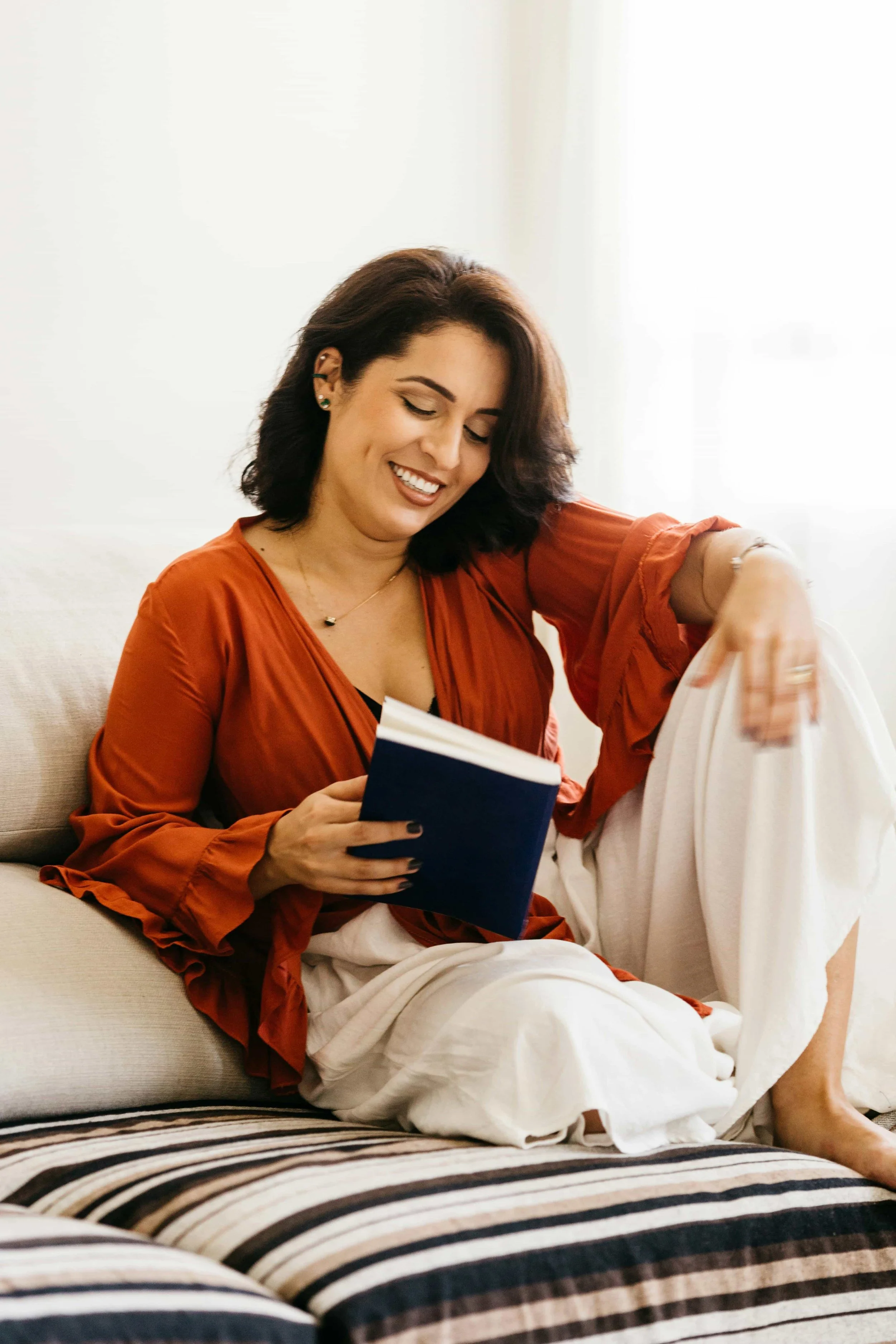 A woman sitting on a sofa, smiling, while reading a blue book. She is wearing a burnt orange blouse and white pants.