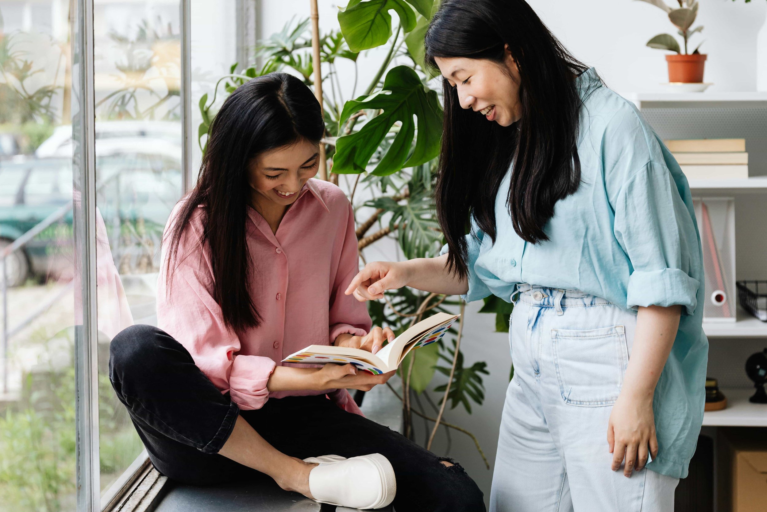 Two women are smiling and looking at a book together inside a room with large windows and green plants.