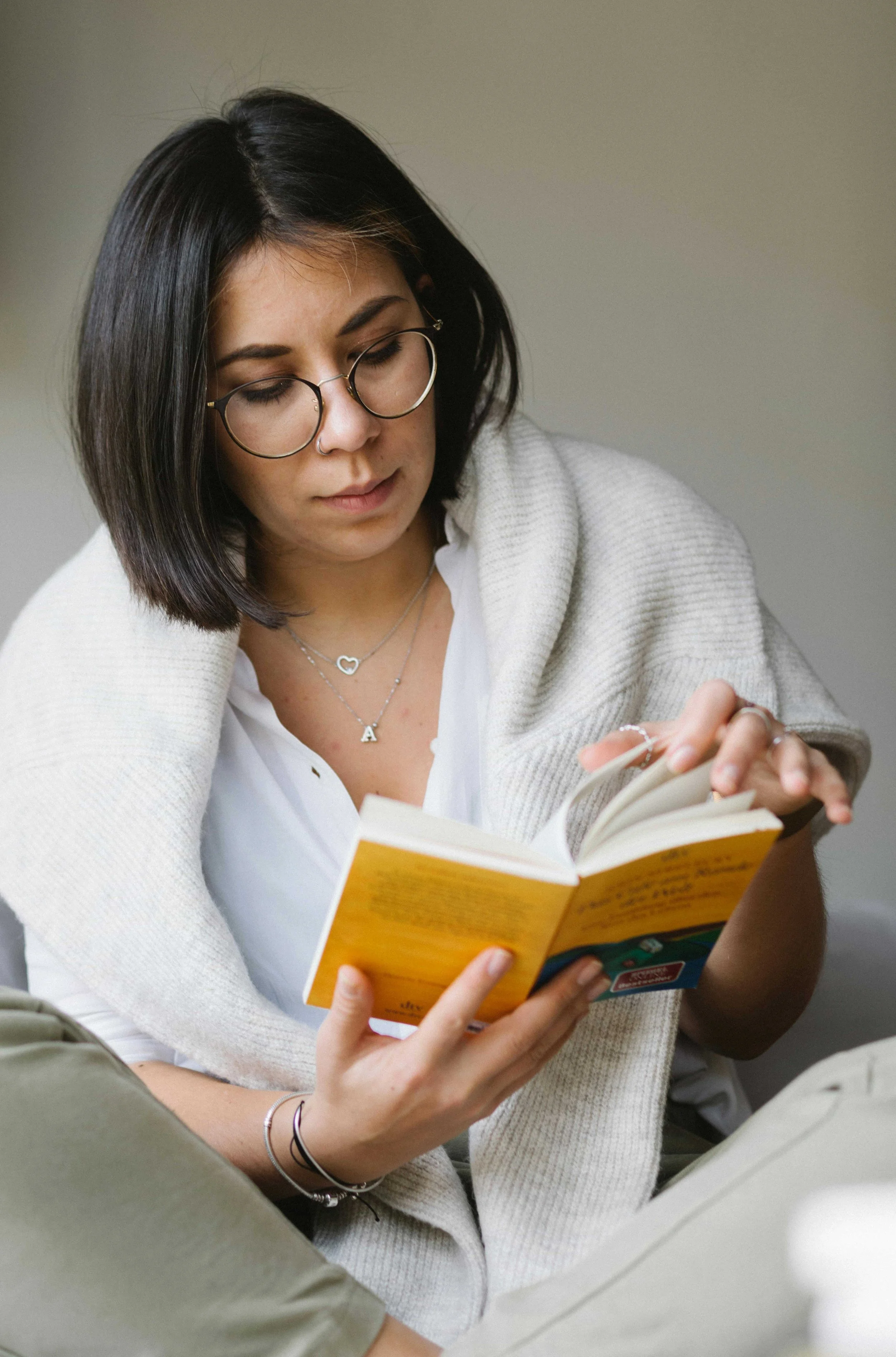 A young woman with black hair, glasses, and a nose ring, is reading a yellow book while seated. She is wearing a white shirt, a light gray cardigan, and layered necklaces with letter and heart pendants.