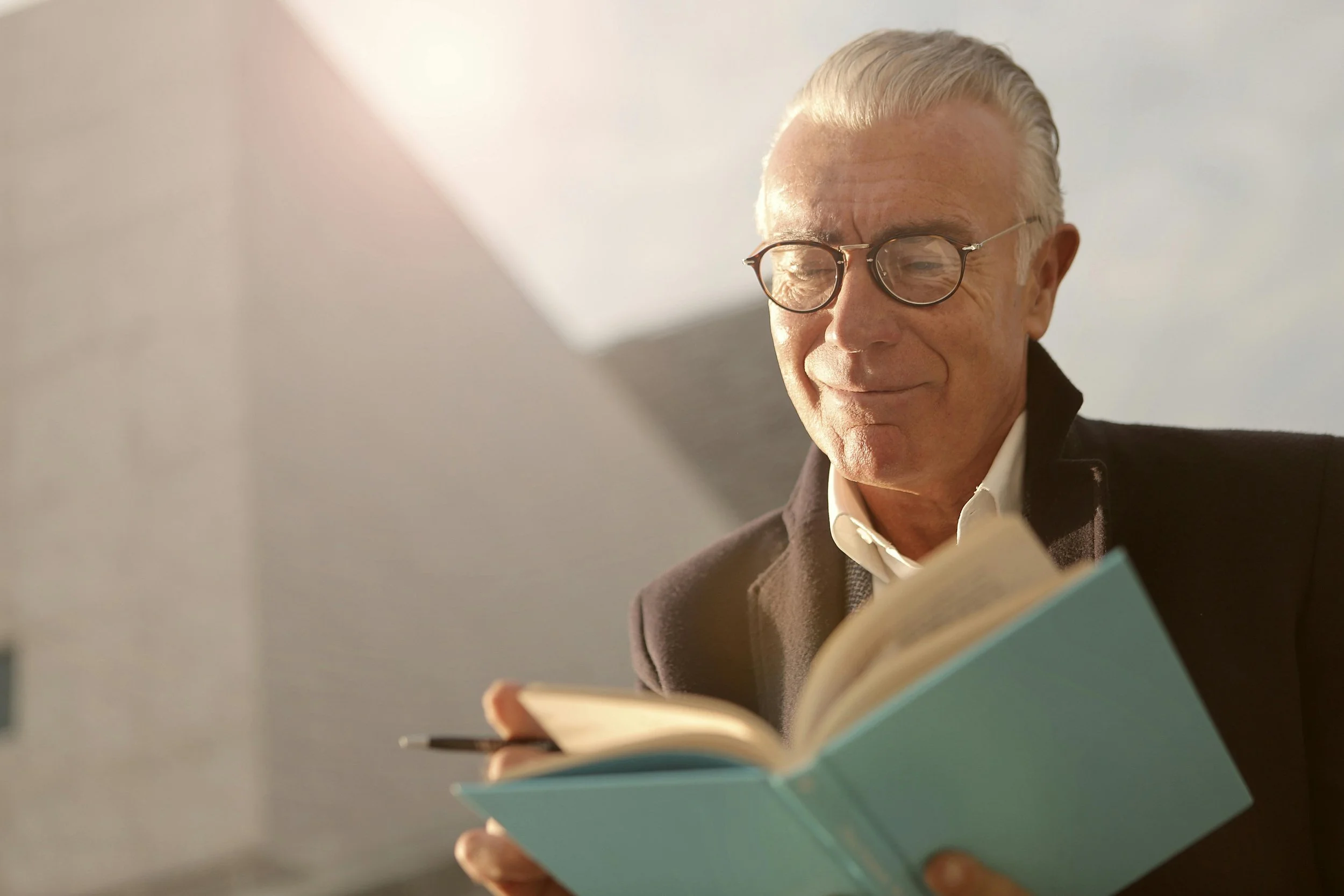 An elderly man with glasses is reading a book and smiling